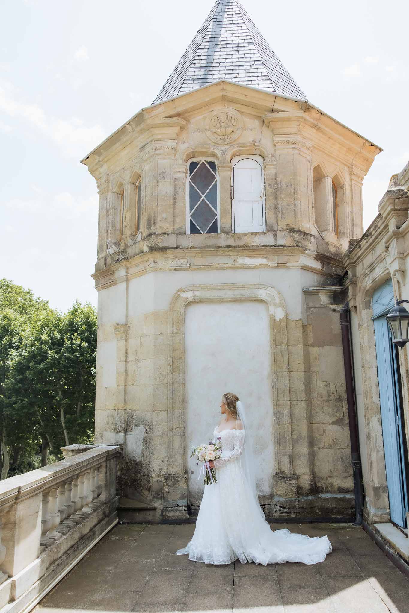 Bride from behind in off-shoulder lace gown with cathedral veil holding blush bouquet before octagonal slate-spired tower