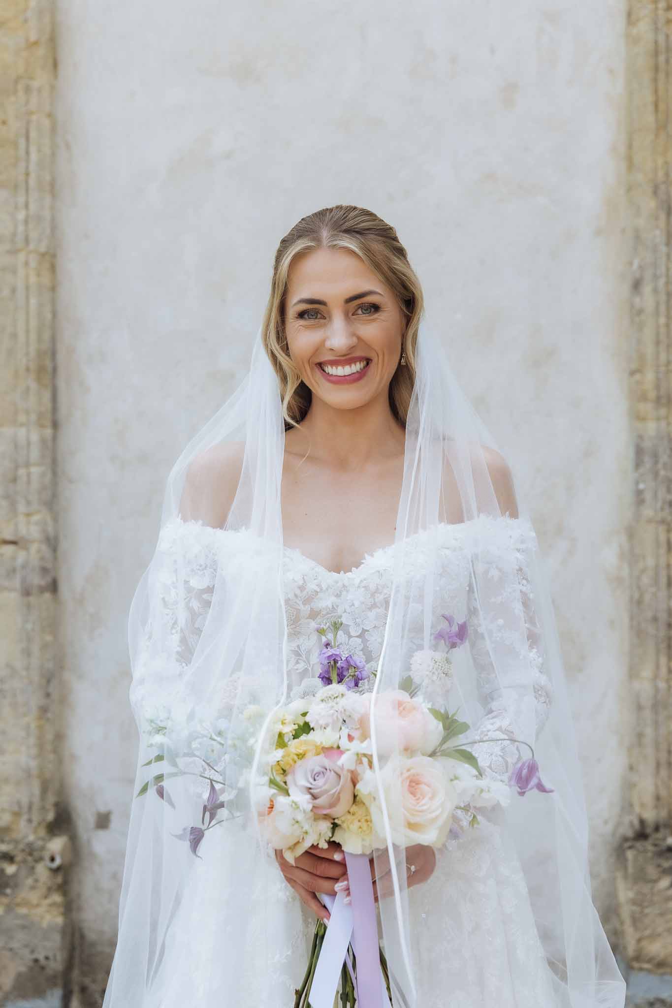 Smiling blonde bride in off-shoulder lace gown holding blush and lavender bouquet with lilac ribbons