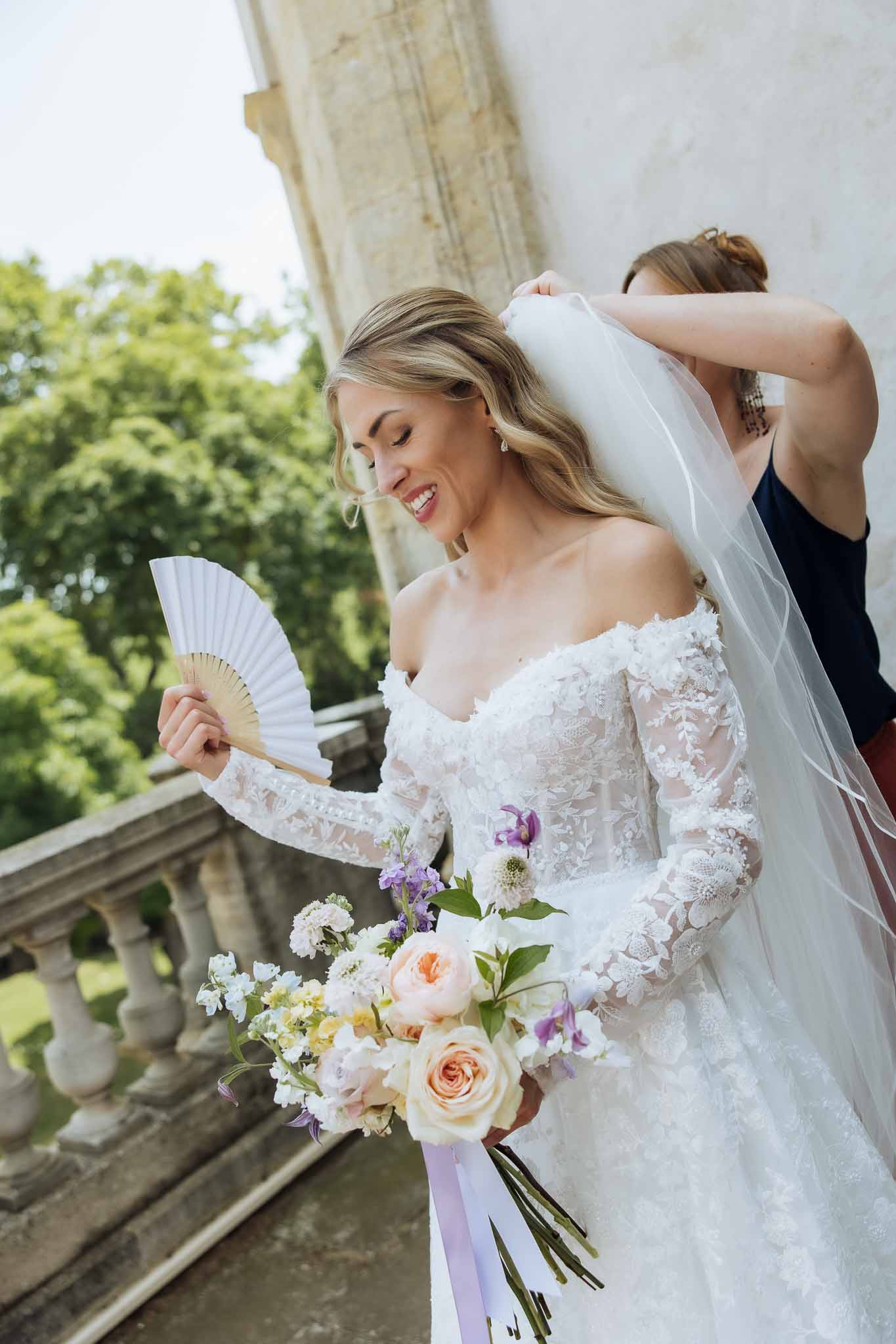 Bridesmaid adjusting bride's veil on stone terrace bride holding peach rose and purple delphinium bouquet