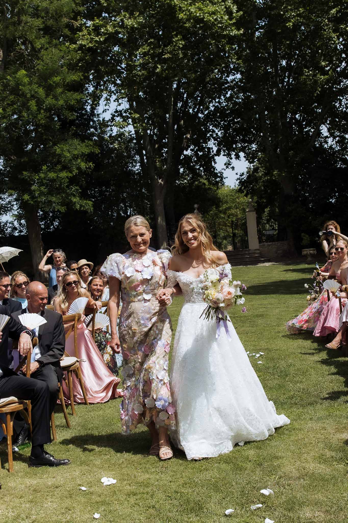 Bride in off-the-shoulder lace ballgown walks down outdoor ceremony aisle holding blush and lavender bouquet accompanied b...