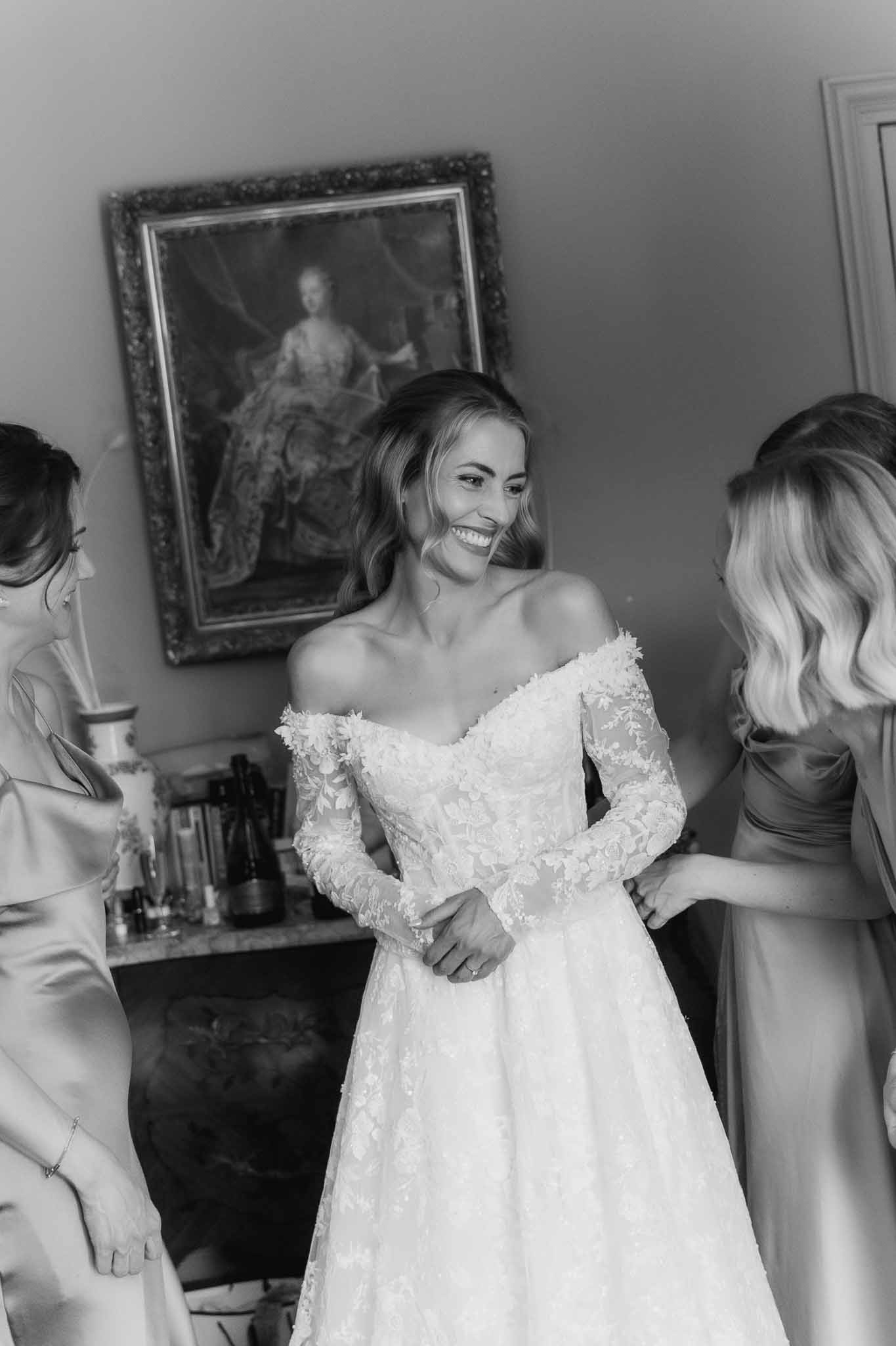Black and white photo of bride smiling as two attendants help her into lace wedding gown in chateau suite
