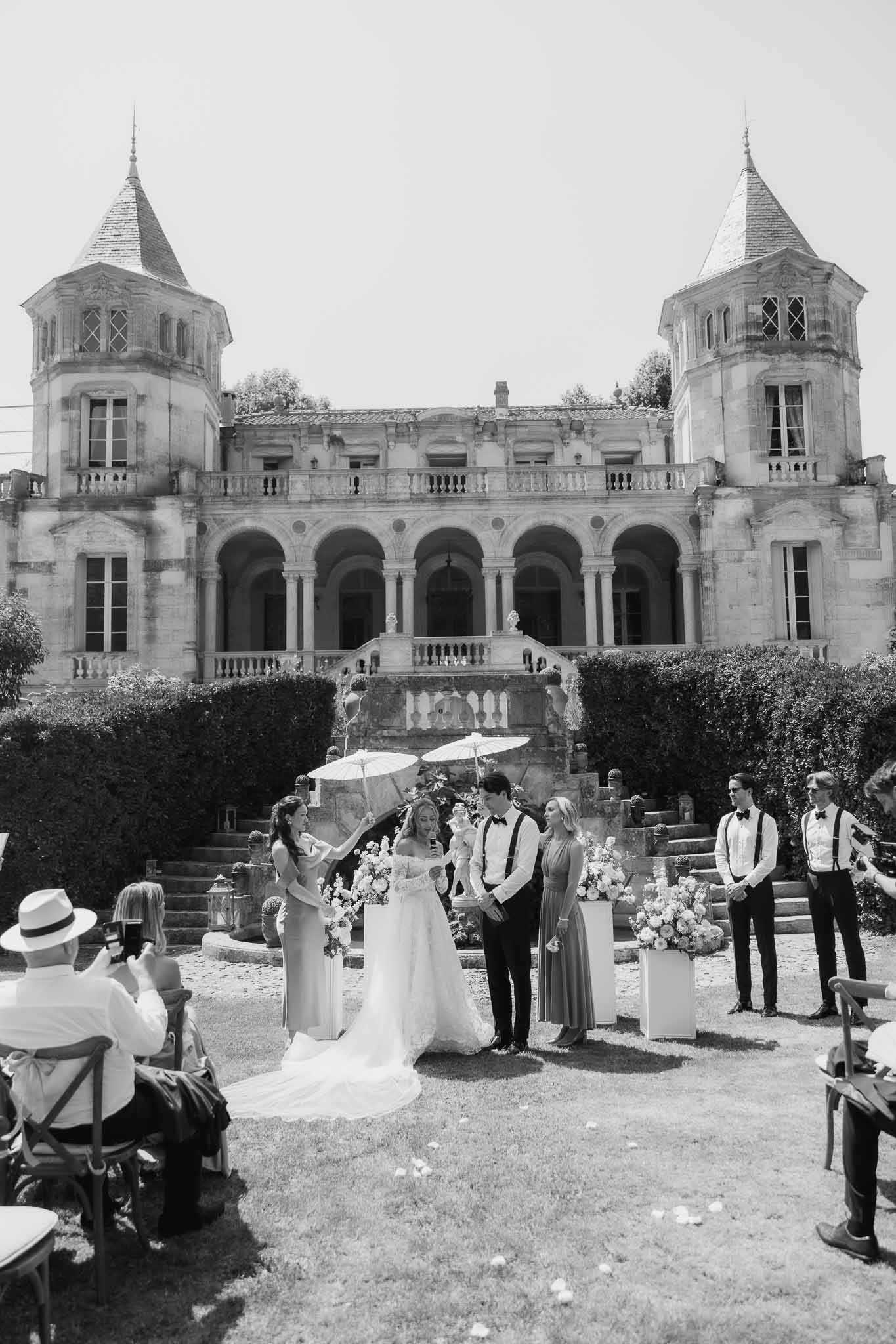 Black and white wide shot of outdoor ceremony before chateau with bridesmaid holding parasol over bride