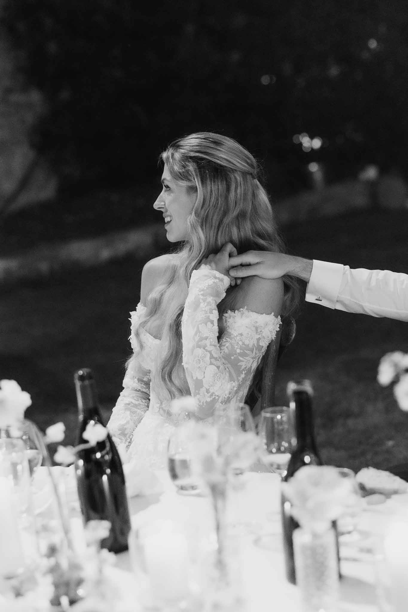 Black and white portrait of bride laughing at reception table with groom's hand on her shoulder during evening dinner