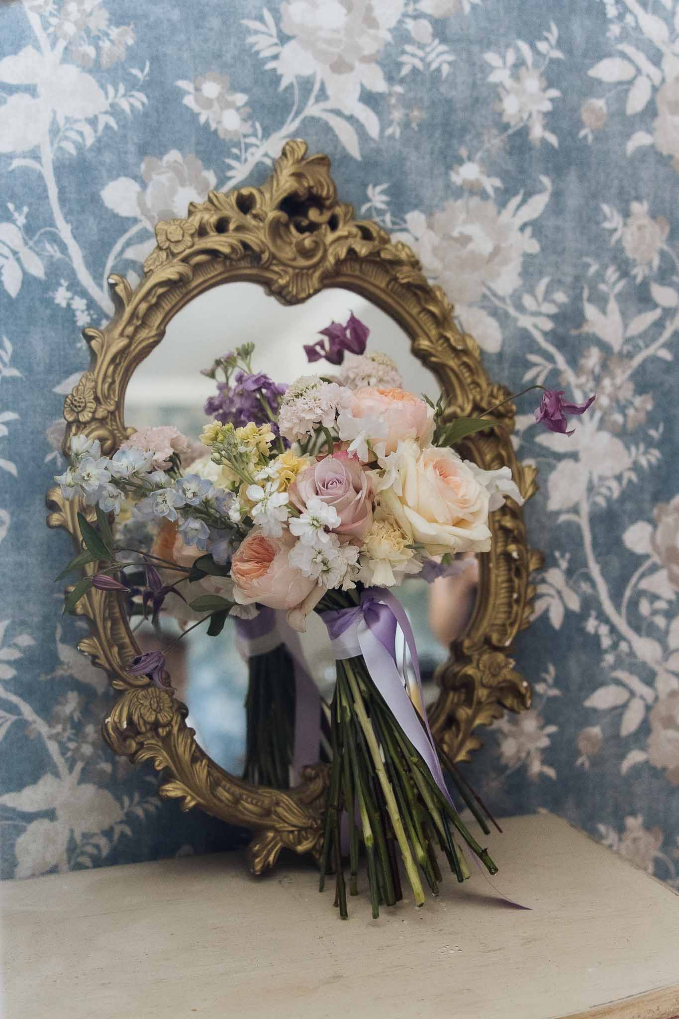Bridal bouquet of peach roses, mauve roses, blue delphinium, and purple sweet peas against gold mirror