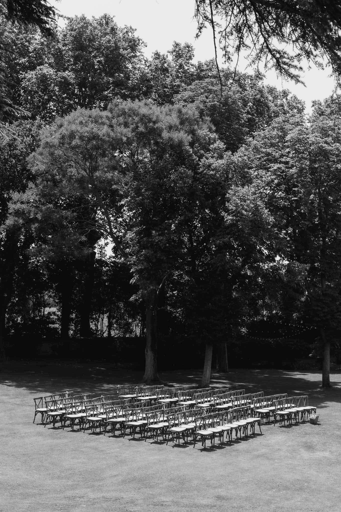 Eighty cross-back chairs in two sections on lawn beneath mature trees before ceremony in elevated B&W shot