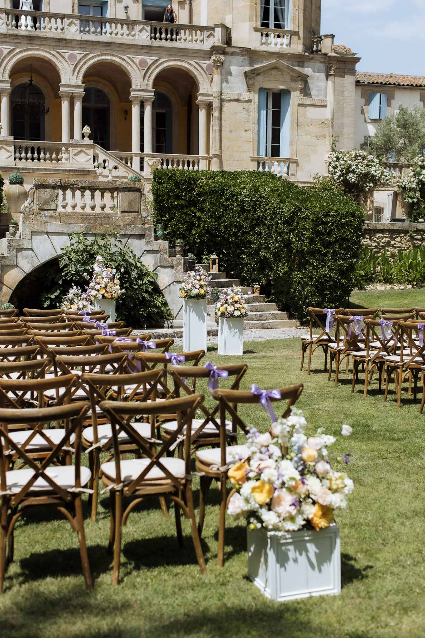 Cross-back chairs with purple bows facing chateau staircase, peach and lilac floral pedestals along aisle