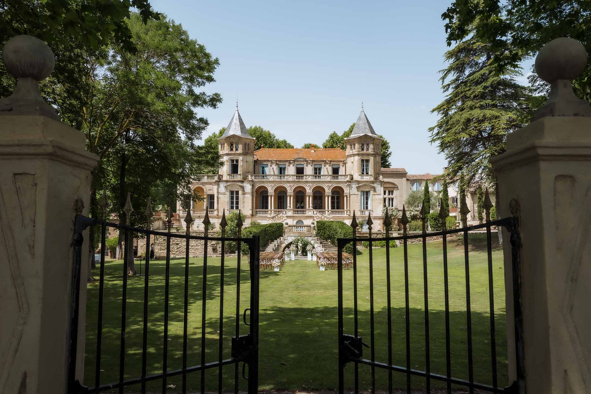 Chateau with conical towers and arcade seen through iron gates with ceremony chairs and floral arch on lawn