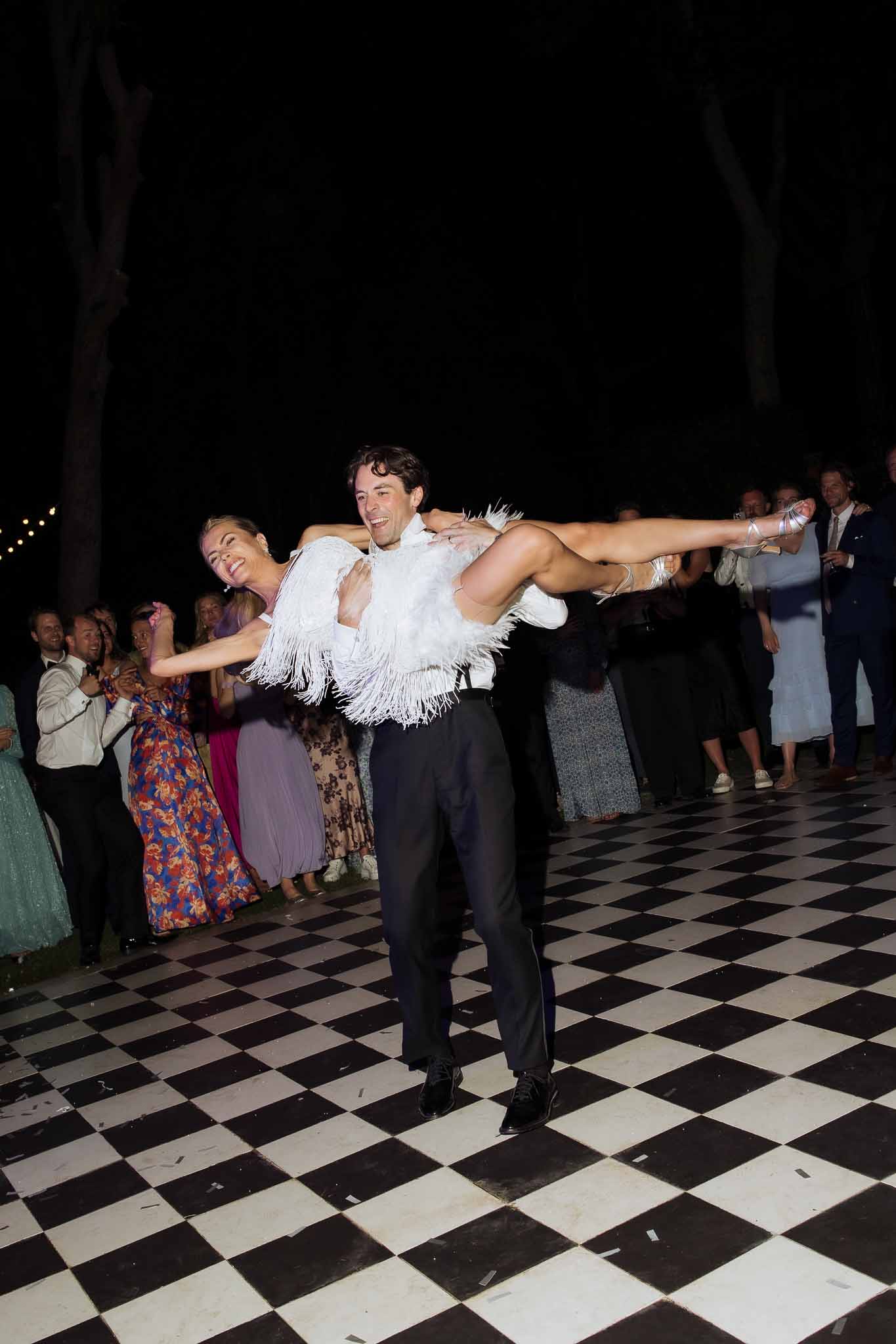 Groom lifting bride during choreographed first dance on checkered outdoor dance floor at evening reception