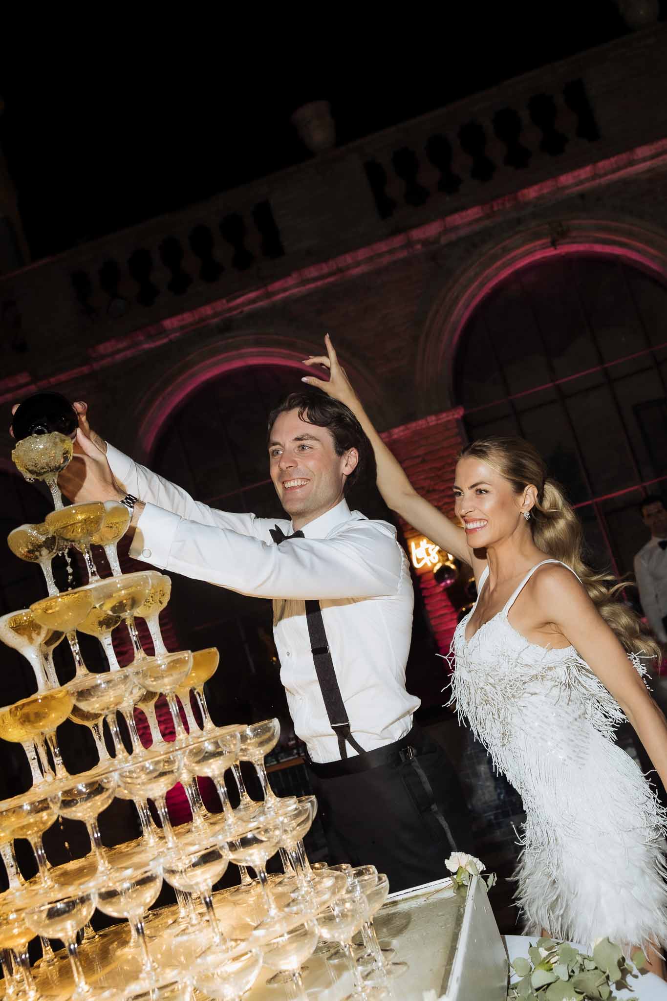 Couple pours champagne into six-tier coupe tower at night with pink and magenta uplighting on stone facade