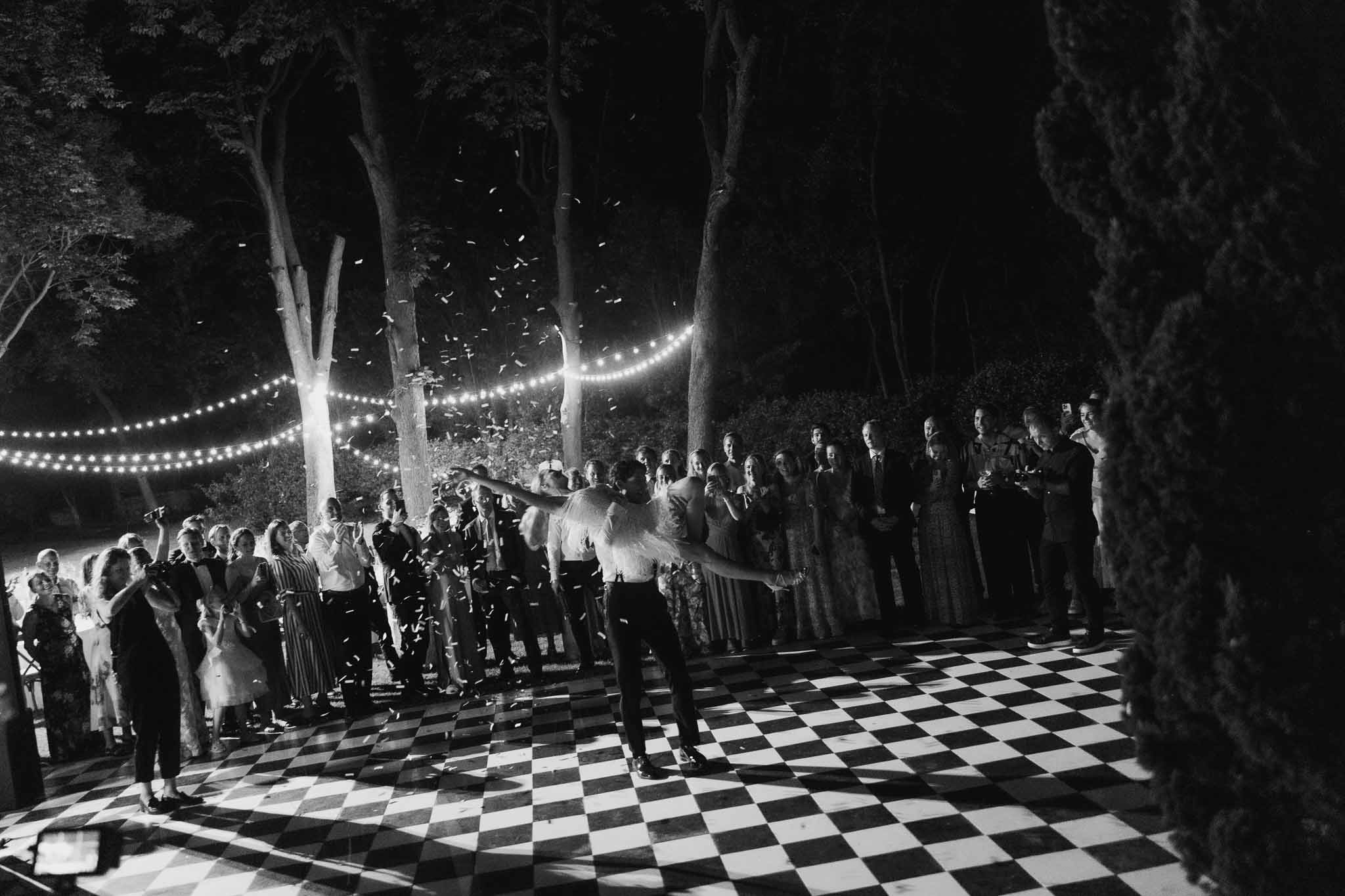 Couple dances on checkered floor with confetti as 45 guests circle under bistro lights in garden in B&W