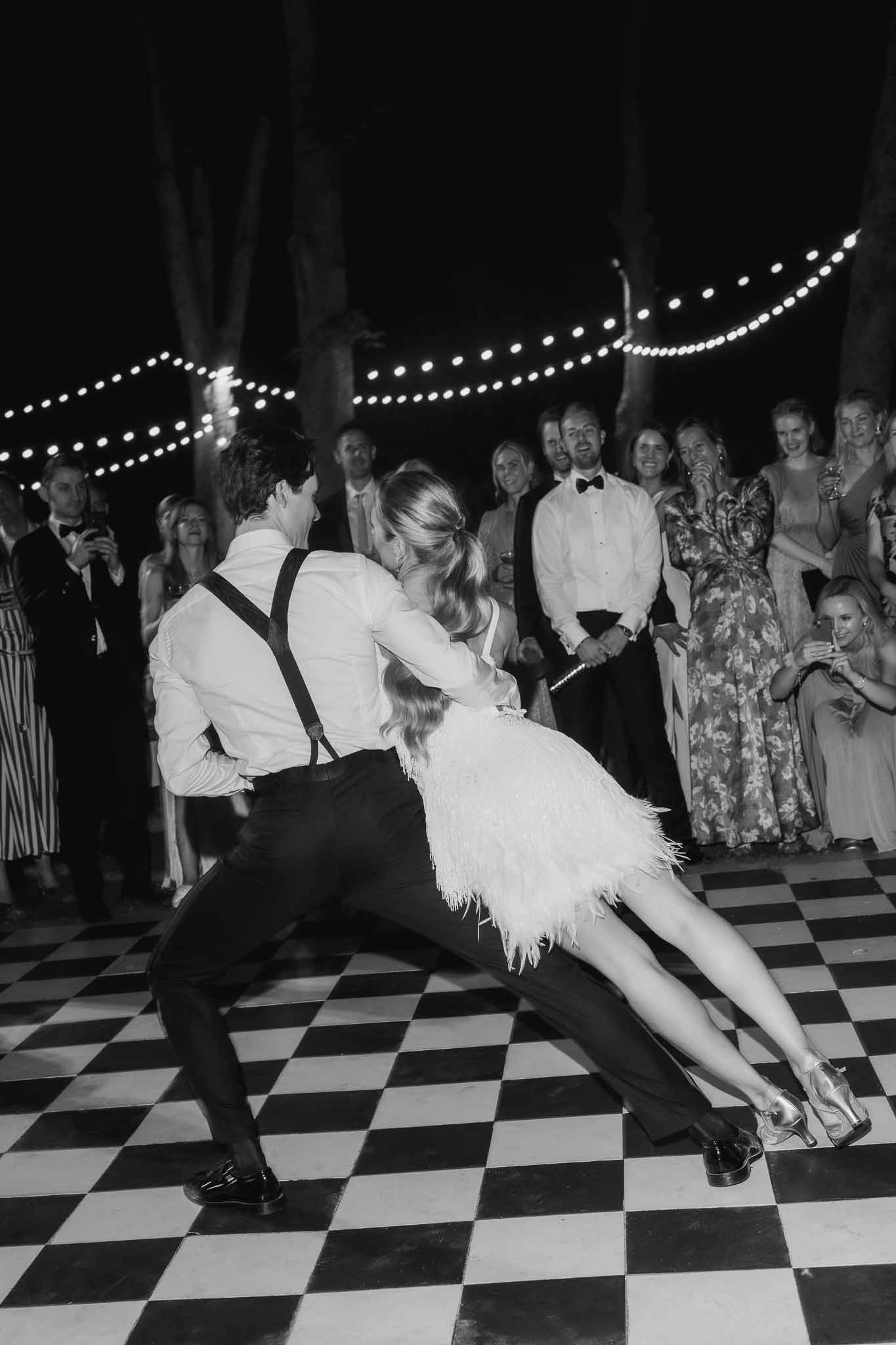 Black and white photo of groom dipping bride on checkered dance floor surrounded by guests under string lights