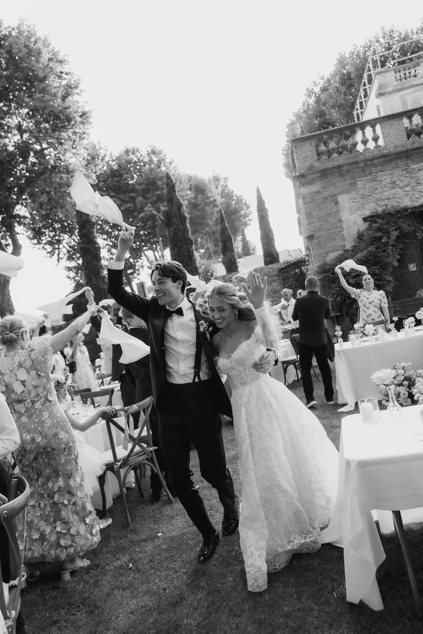 Couple entering outdoor reception as guests wave white napkins on chateau lawn in black and white