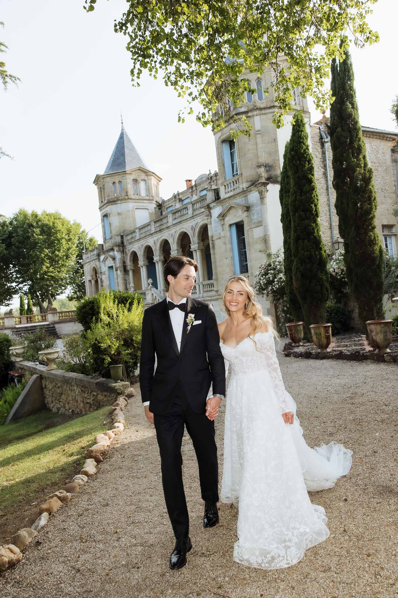Couple walking toward camera at chateau with colonnades, turret, and cypress tree backdrop