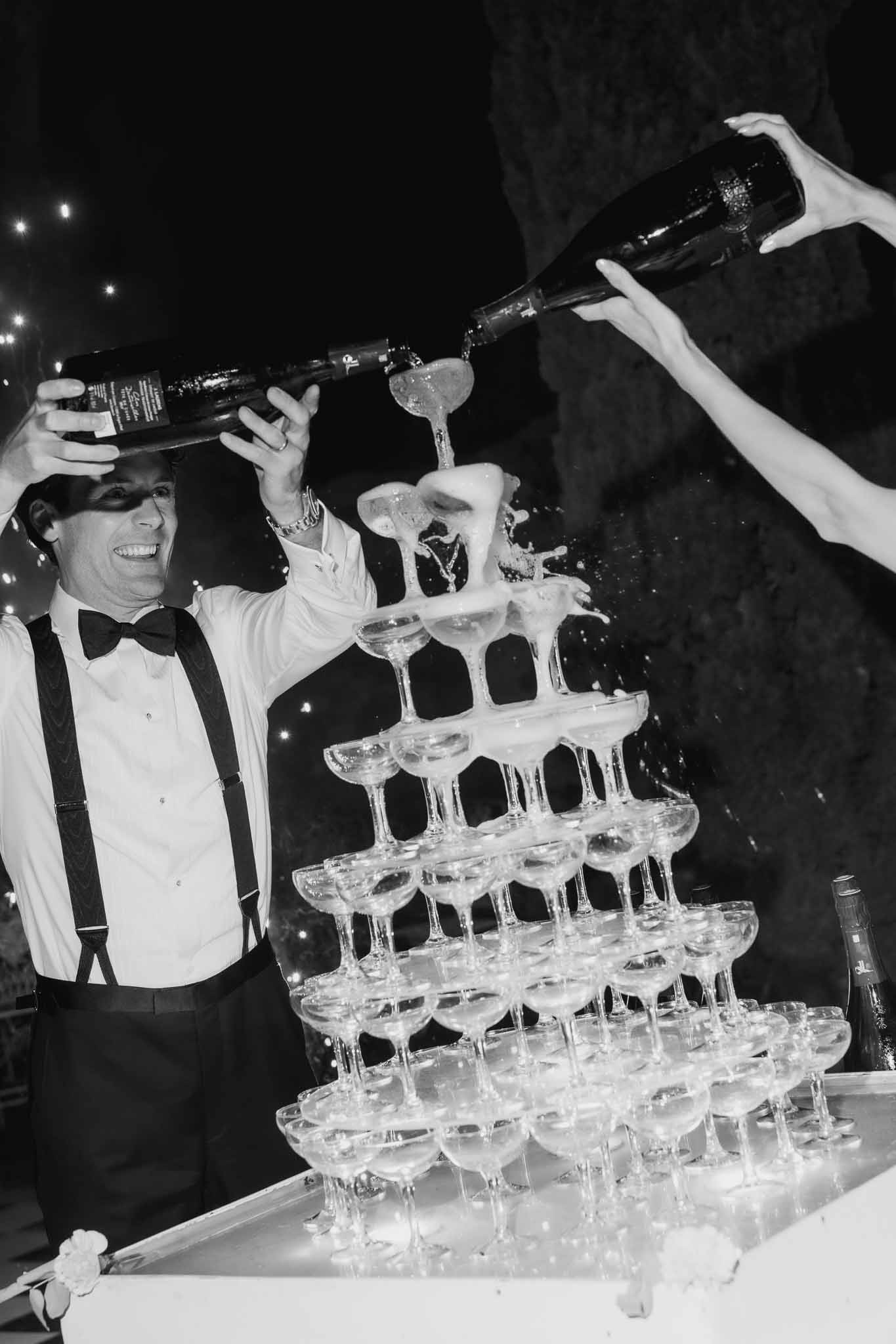 Black and white of champagne tower pour with foam cascading through coupe pyramid under string lights at night