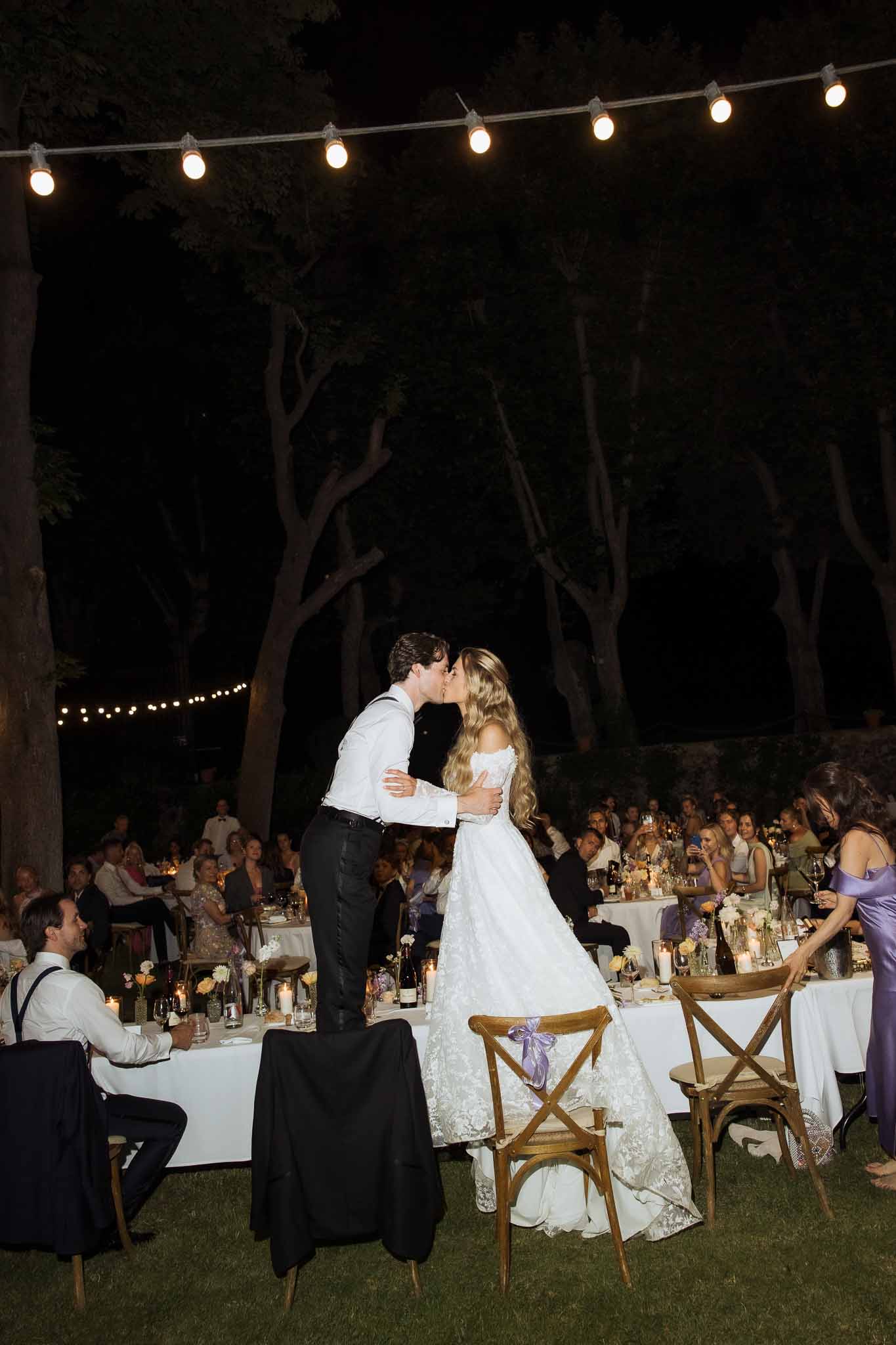 Couple kissing between banquet tables with lavender ribbon chairs under Edison lights at night reception