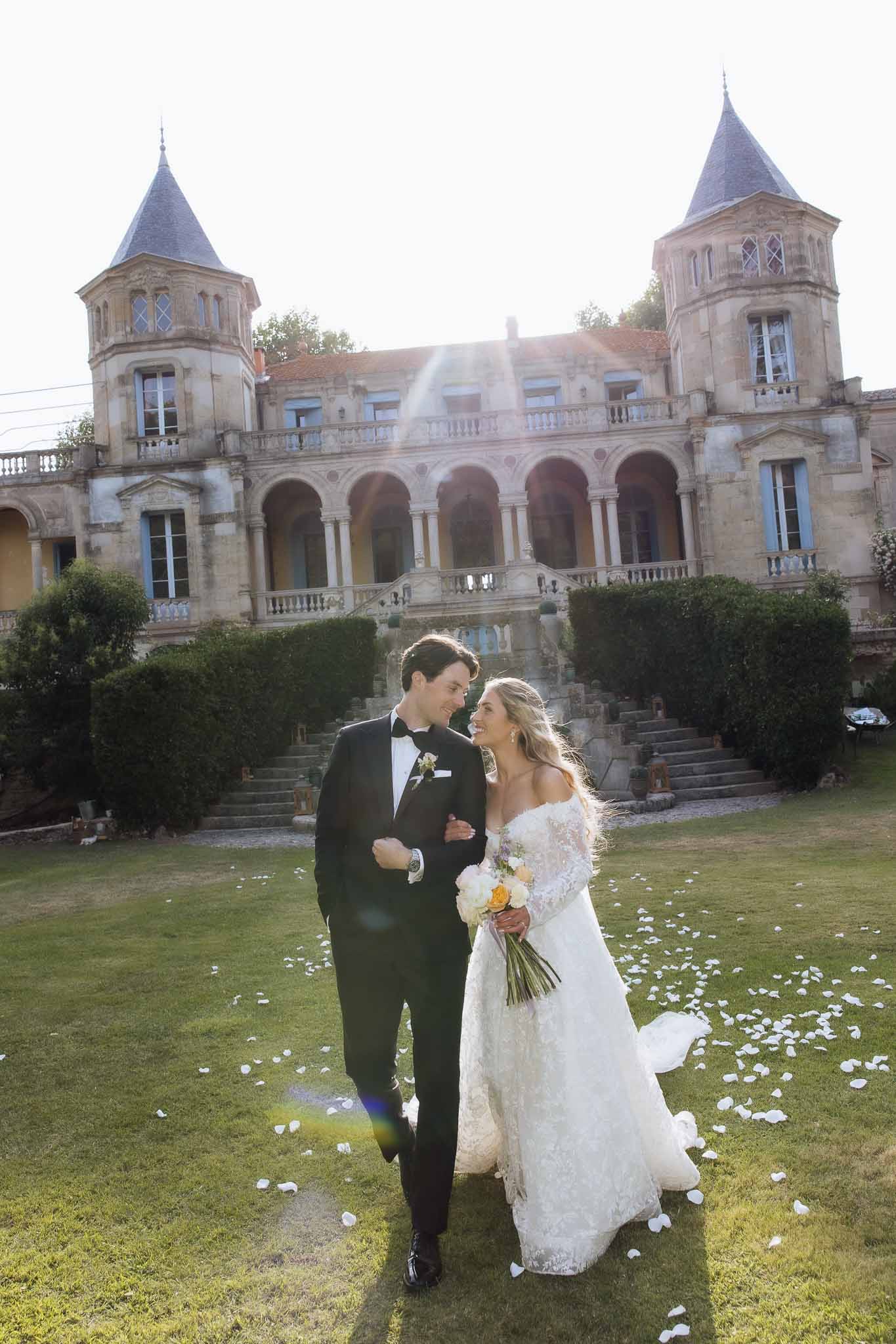 Bride and groom with sun flare on chateau lawn with turreted stone building and scattered rose petals