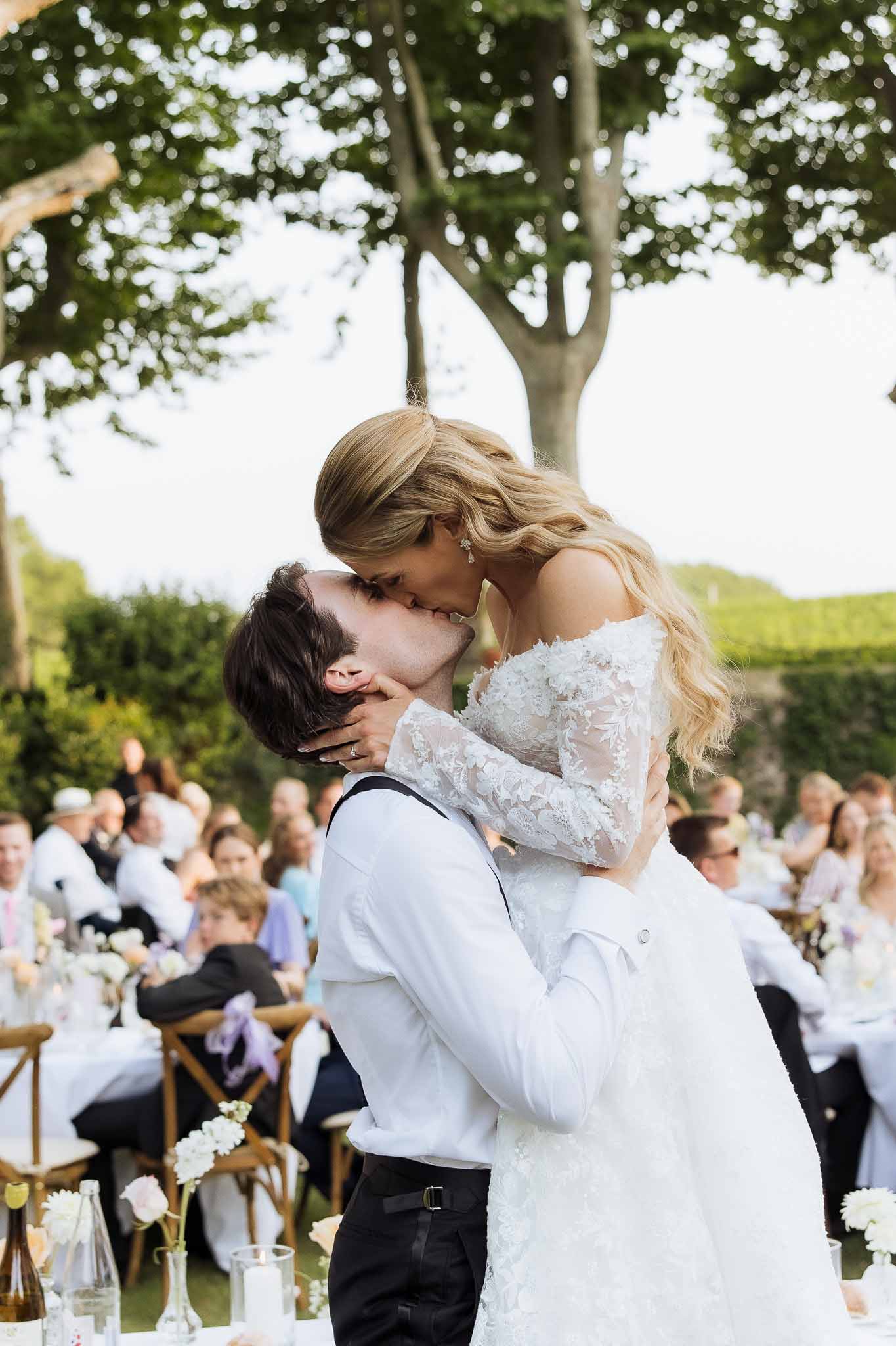 Groom lifts bride for a kiss during an outdoor garden reception with banquet tables and guests in the background