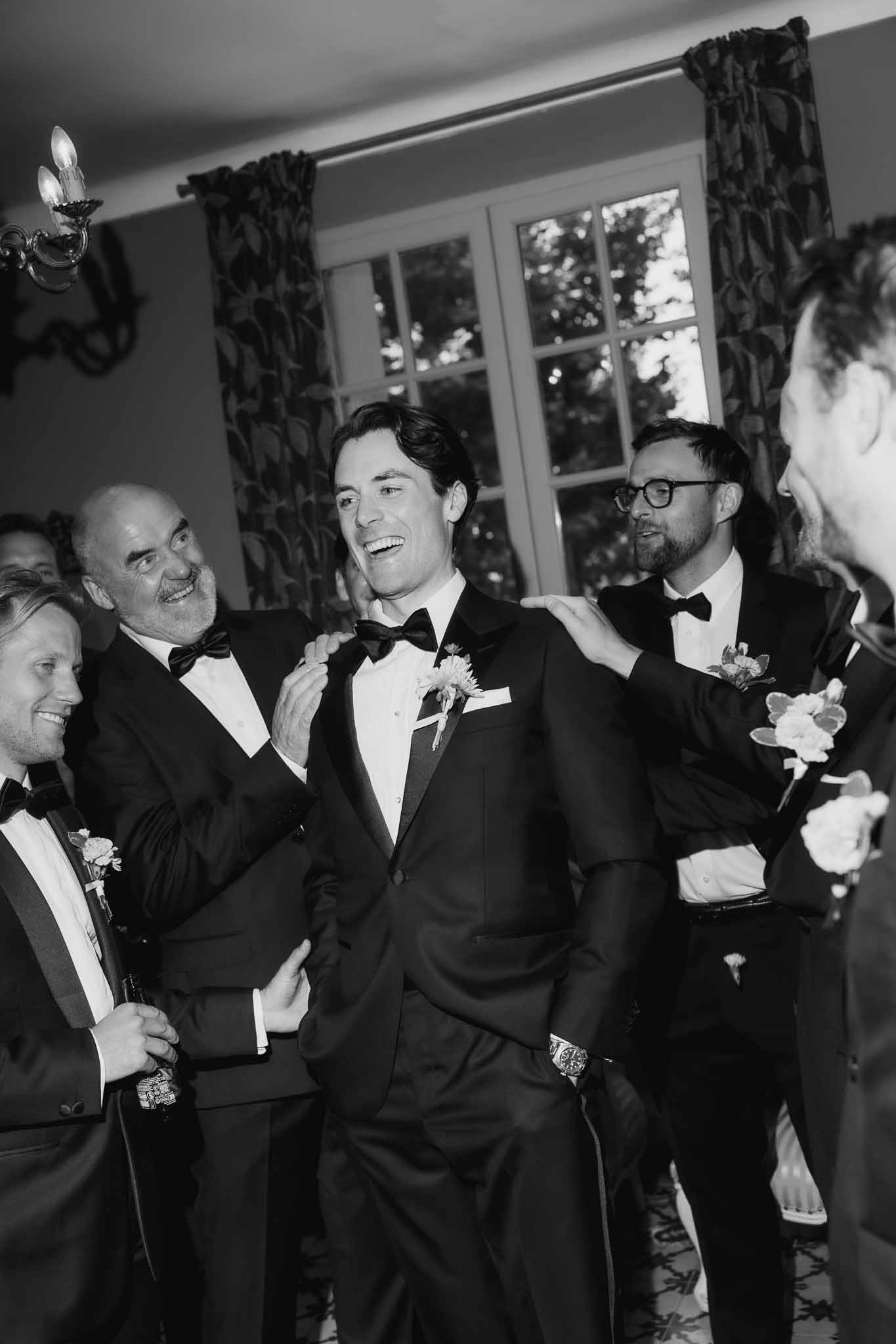 Black and white of groomsmen adjusting groom's bow tie and jacket while laughing in chateau room
