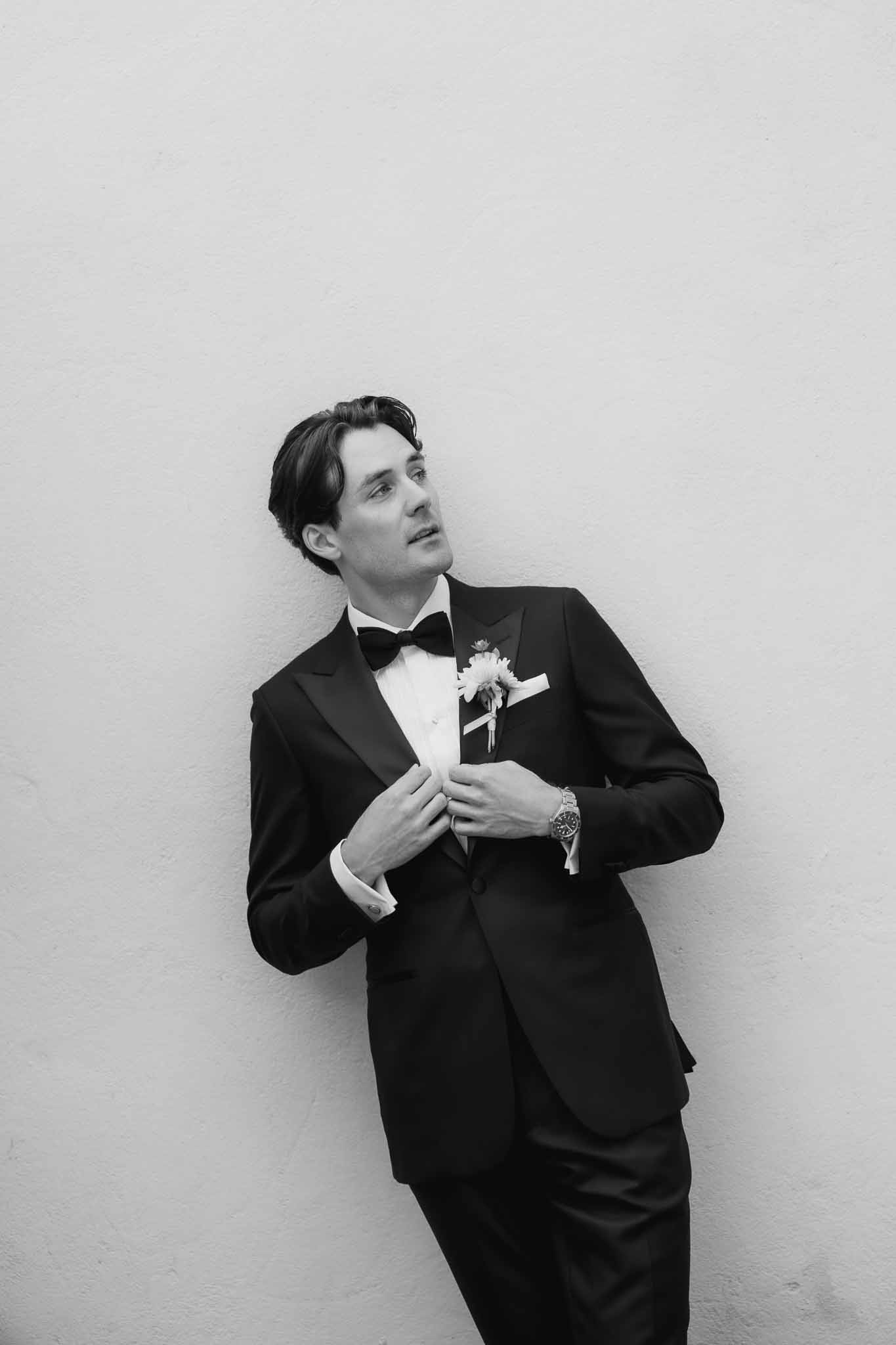 Black-and-white portrait of groom in black tuxedo and bow tie adjusting jacket against plain wall