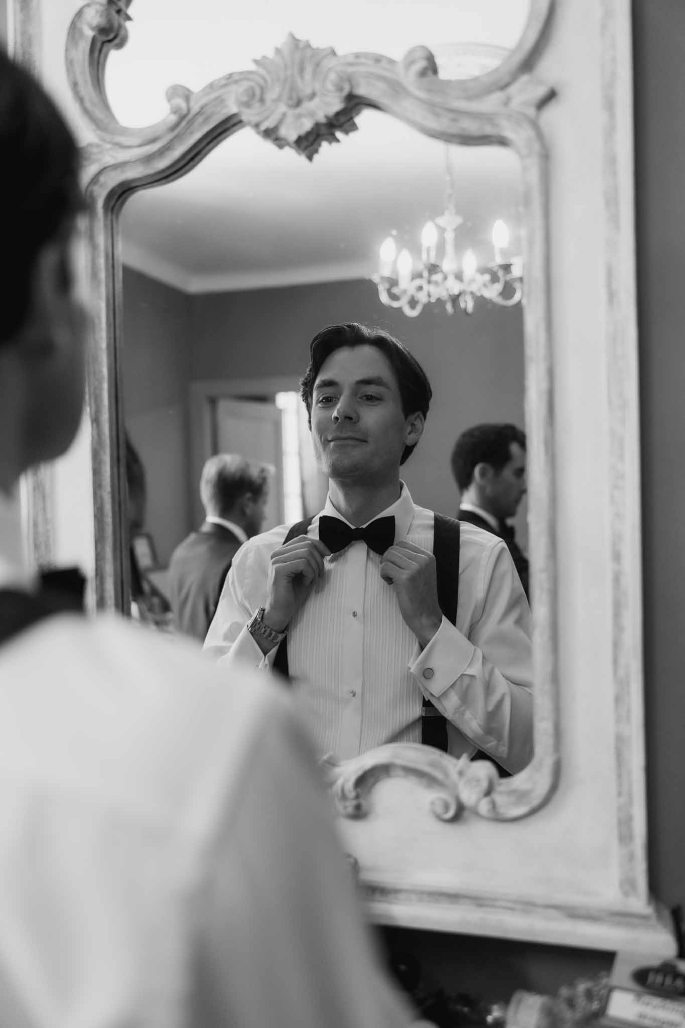Black-and-white image of groom adjusting bow tie in ornate French rococo mirror with groomsmen visible in reflection