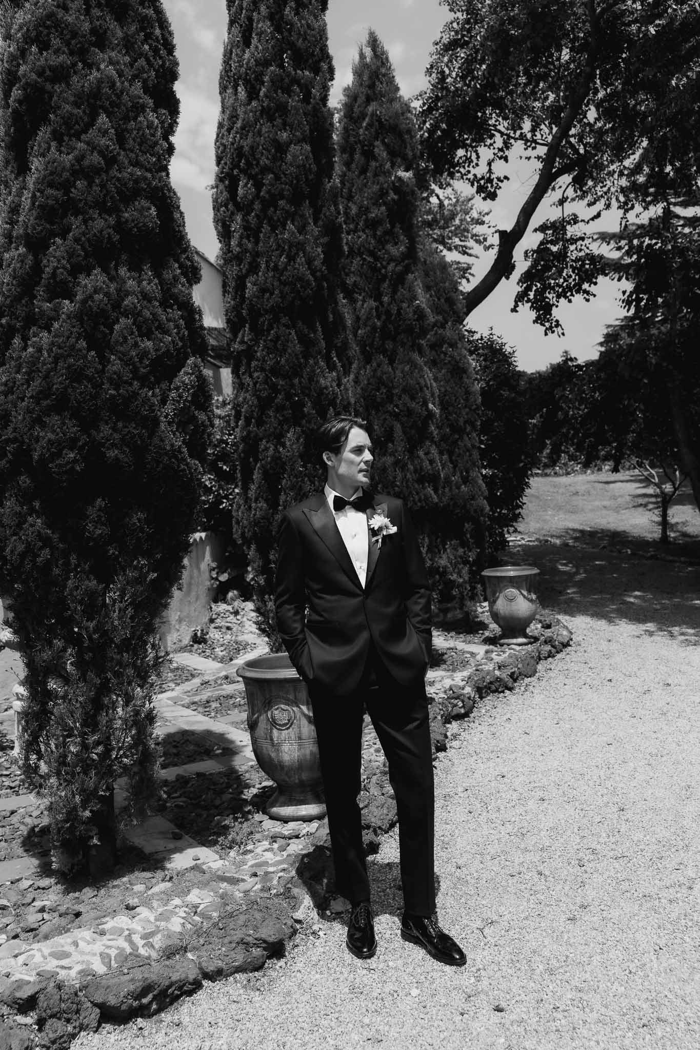 Black and white groom portrait in tuxedo with hands in pockets on gravel path with cypress trees