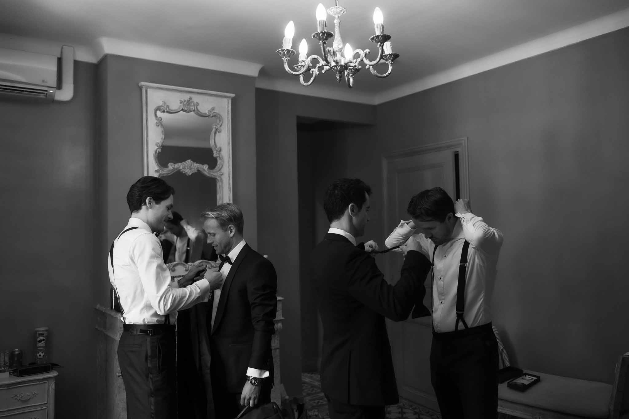 Black and white scene of four groomsmen helping each other with tuxedos and bow ties in ornate chateau room