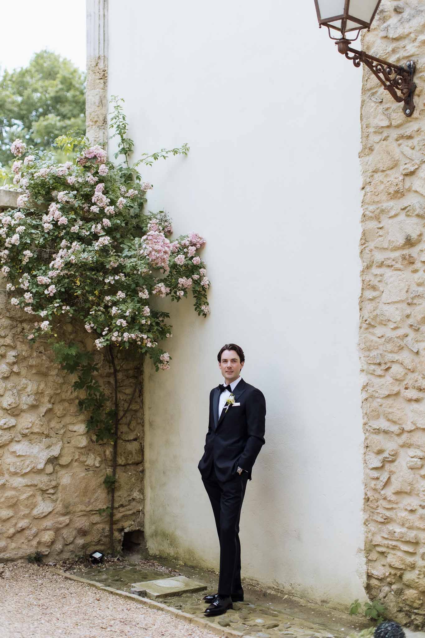 Groom in black tuxedo standing in French chateau courtyard with climbing blush roses on stone wall