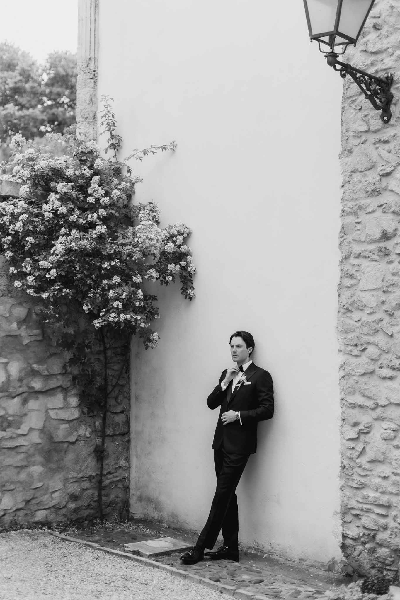 Black and white full-length portrait of groom in dark suit and bow tie leaning against a stone wall at a French estate