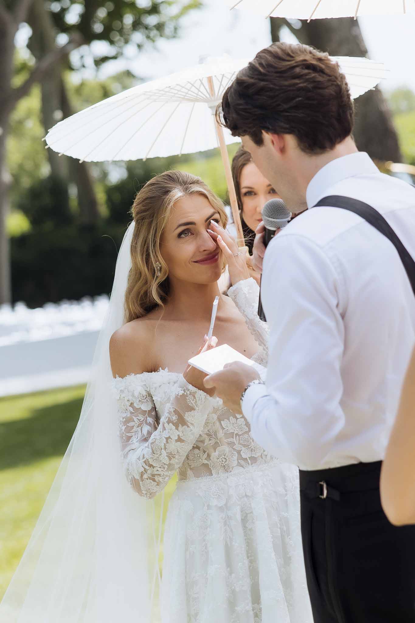 Bride wipes tear as groom reads vows during outdoor ceremony under white paper parasol on estate lawn