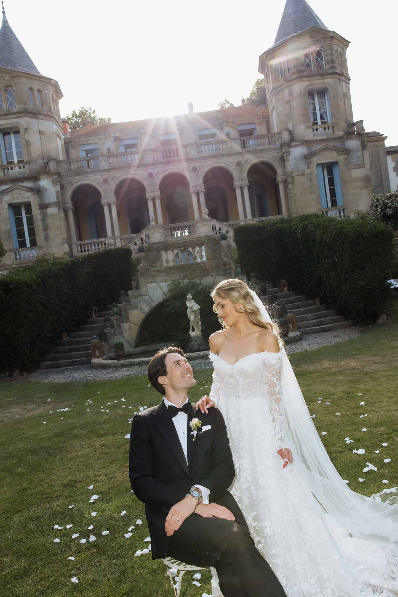 Groom seated on lawn gazing up at bride in off-shoulder lace gown before chateau with twin turrets