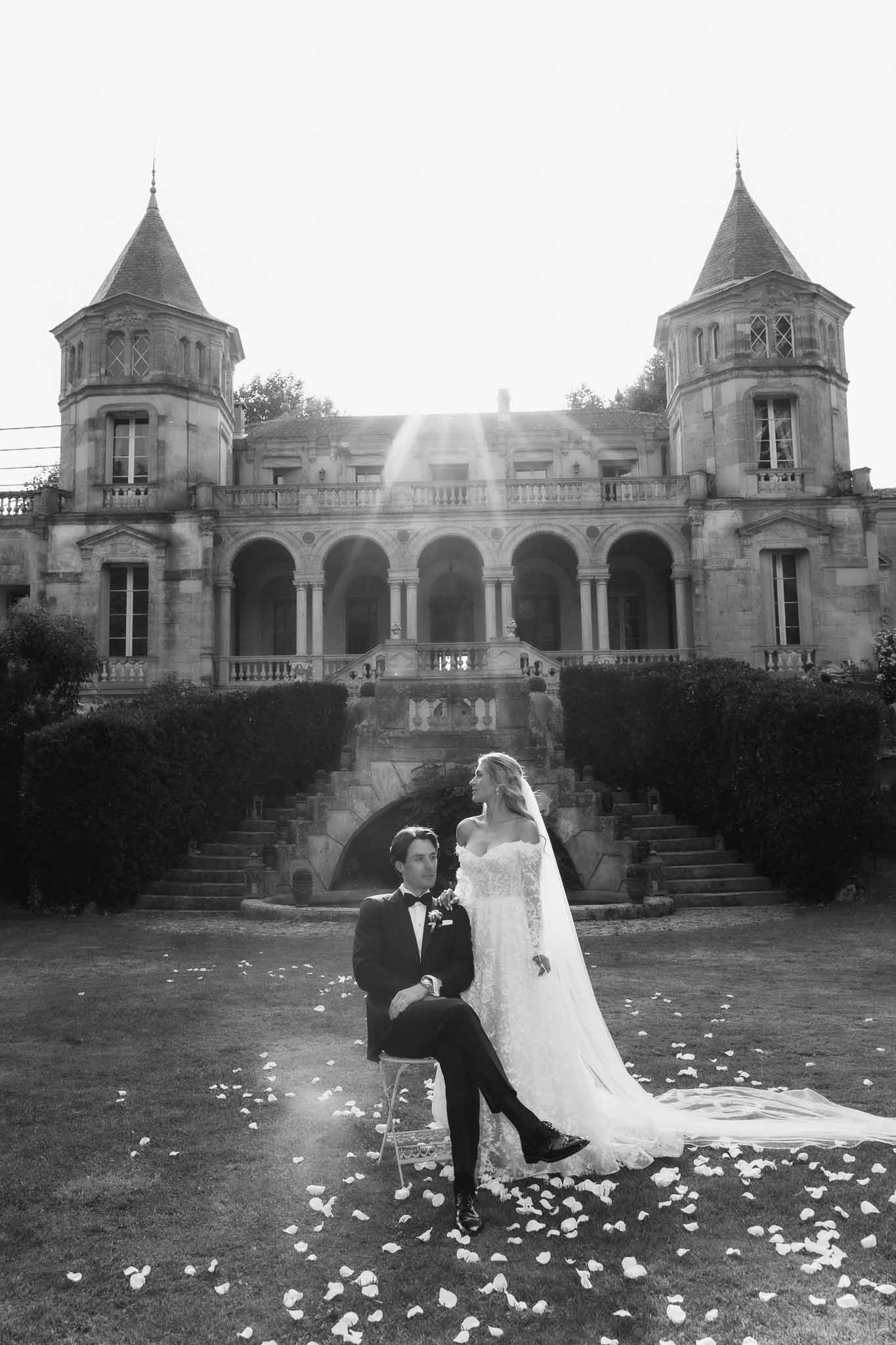 Black and white couple before twin-towered chateau with cathedral veil fanned on lawn and lens flare