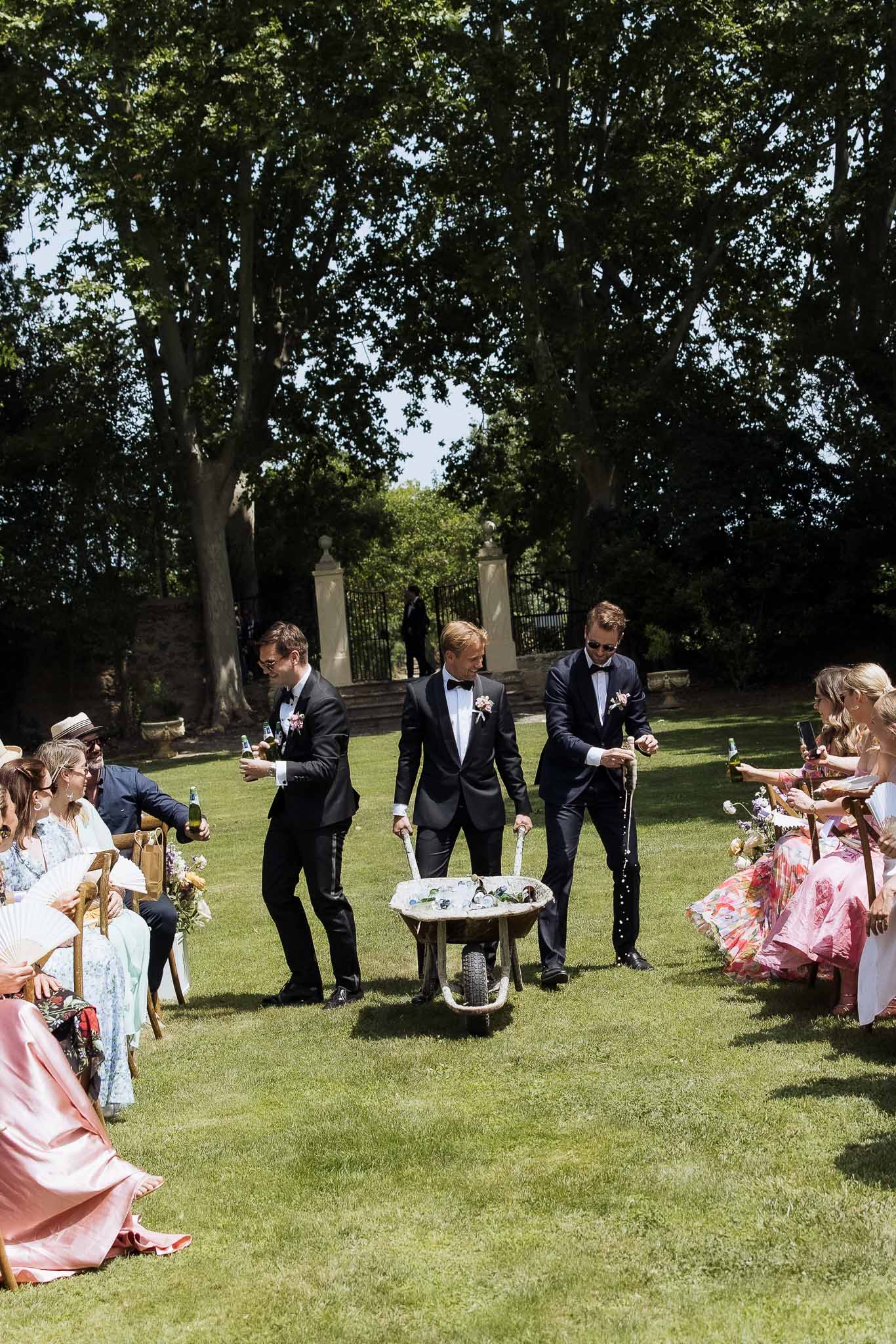 Three groomsmen distributing champagne from wheelbarrow to seated guests in formal garden