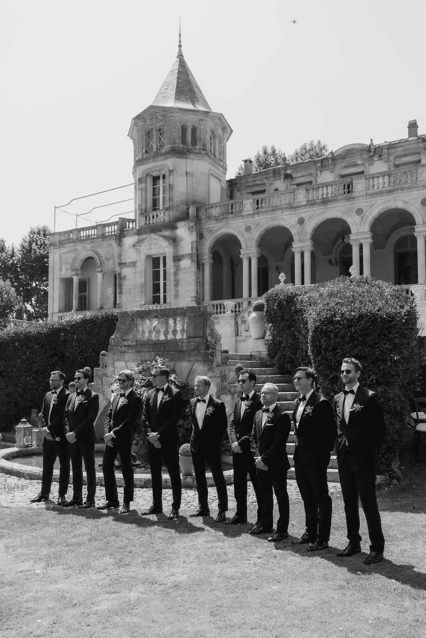 Black-and-white group portrait of groom and eight groomsmen in tuxedos on French chateau steps