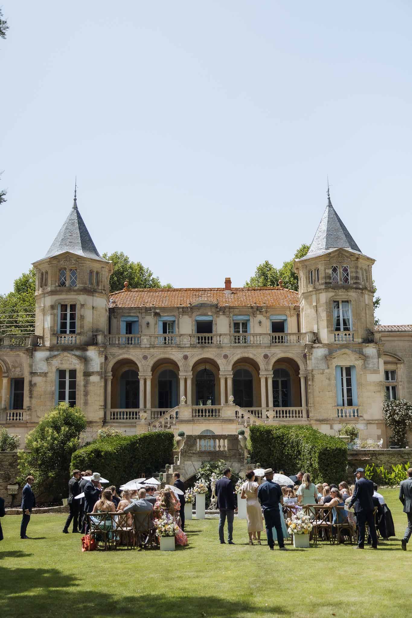 35 guests with parasols mingling on lawn before twin-towered chateau with floral cube planters