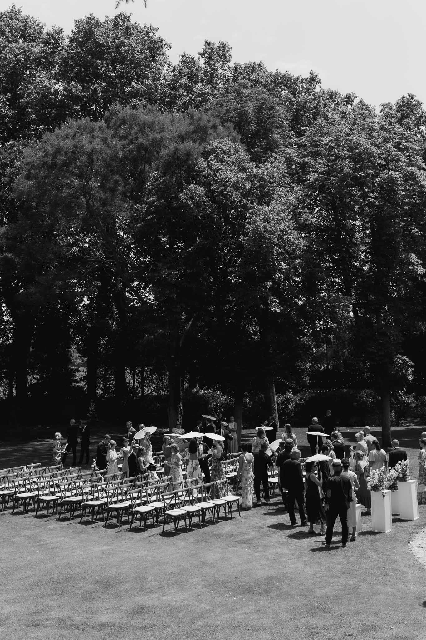 Guests arriving at outdoor ceremony with crossback chairs, white parasols, and pedestal florals in B&W