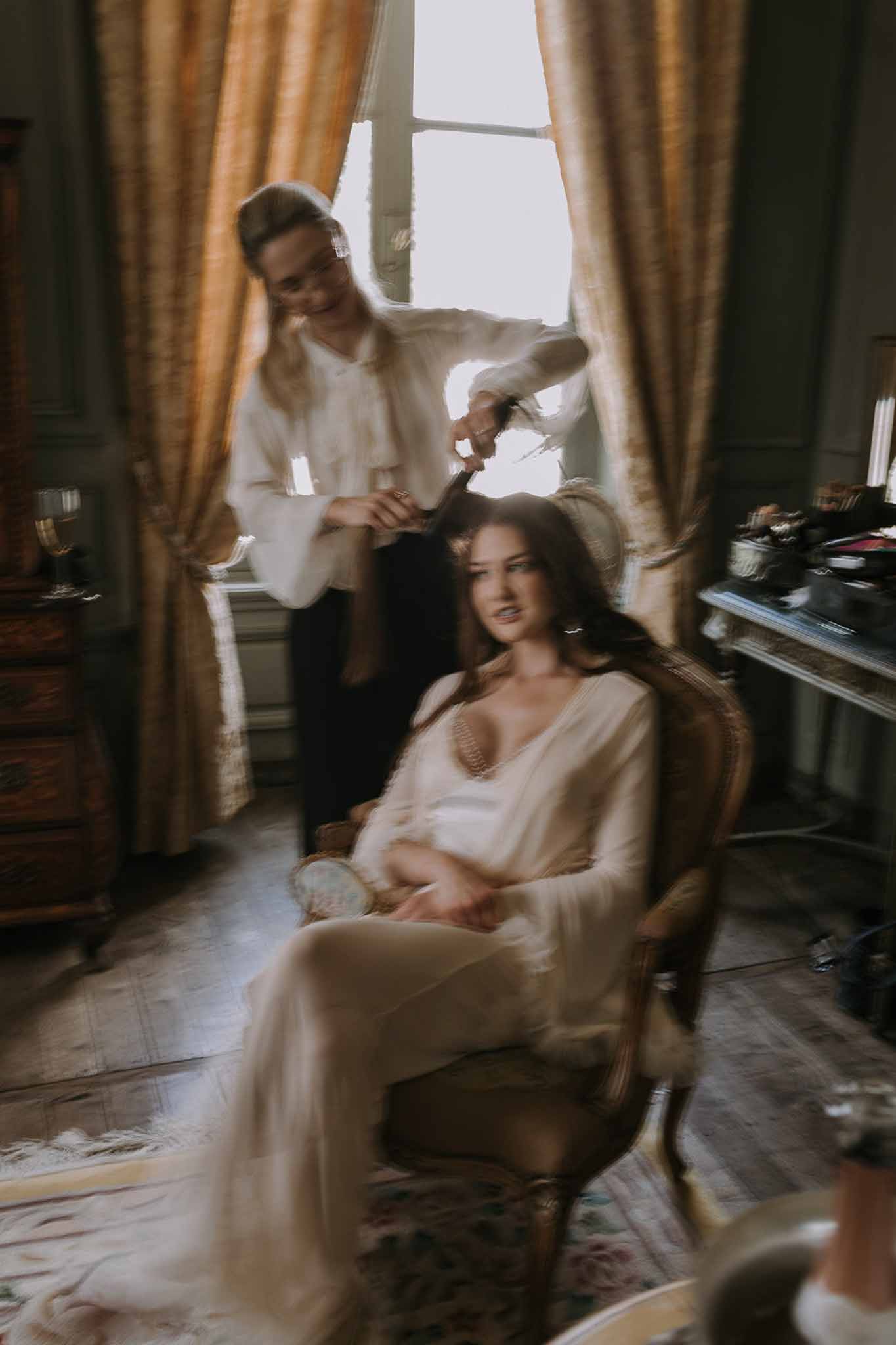 Motion-blurred getting ready scene of hairstylist working on bride in ivory robe in gold-curtained chateau room