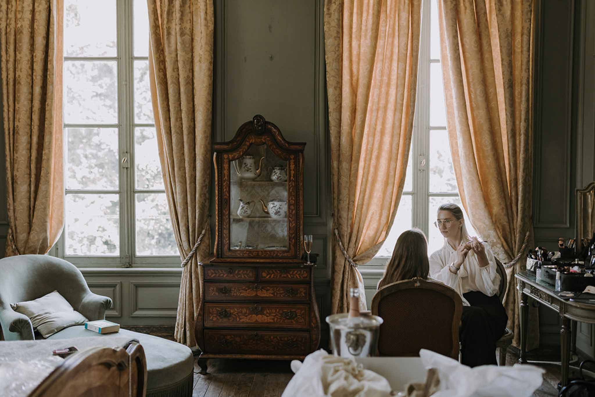 Bride having makeup done in sage-panelled chateau room with gold curtains and marquetry armoire