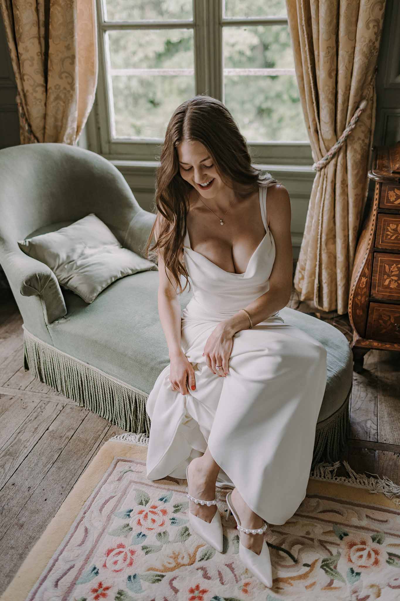 Bride adjusting pearl-strap heels on sage velvet chaise longue in French chateau dressing room