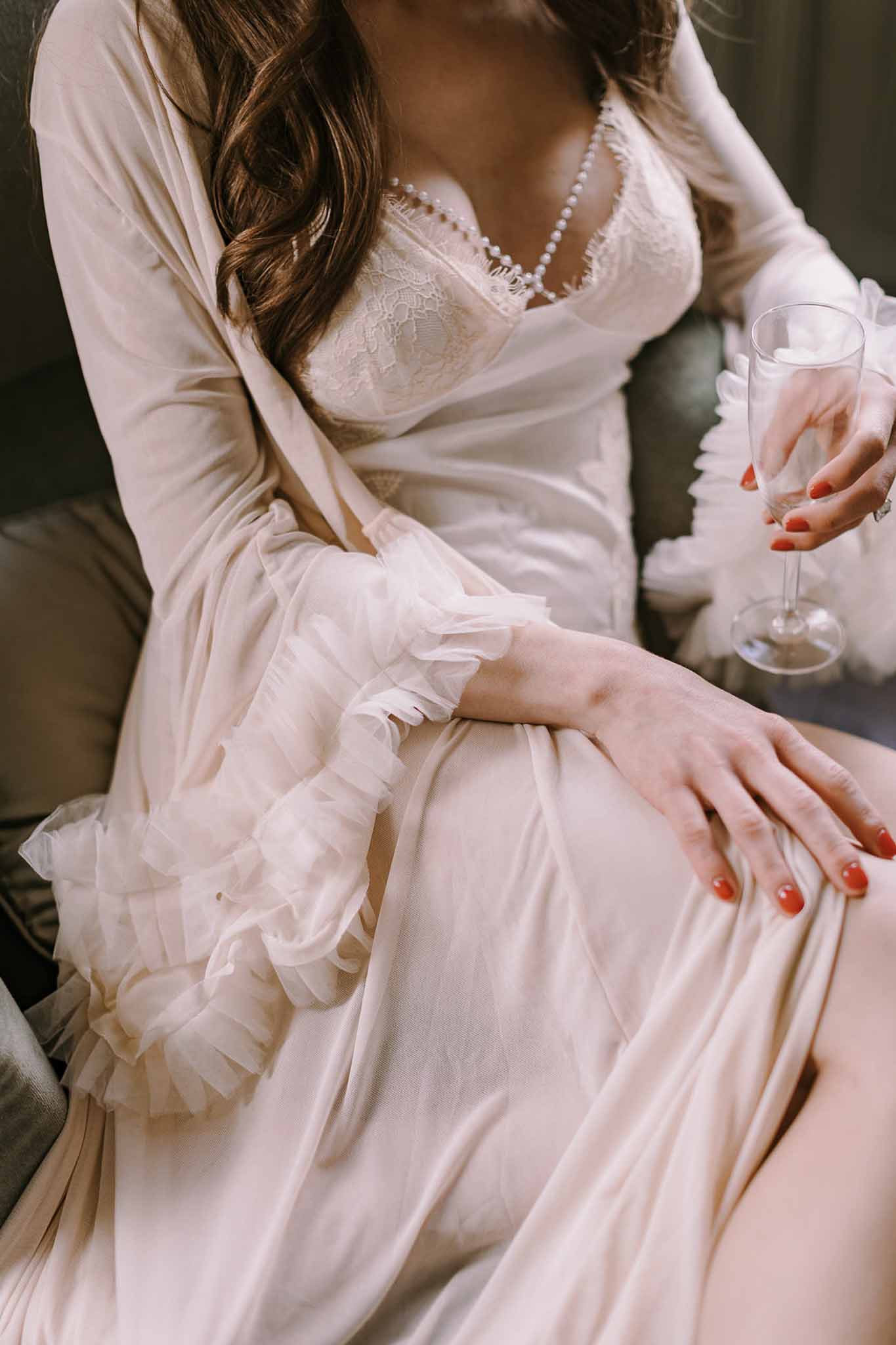 Bride in cream lace bodice gown and ruffled ivory robe holding champagne with red-orange nails on dark sofa