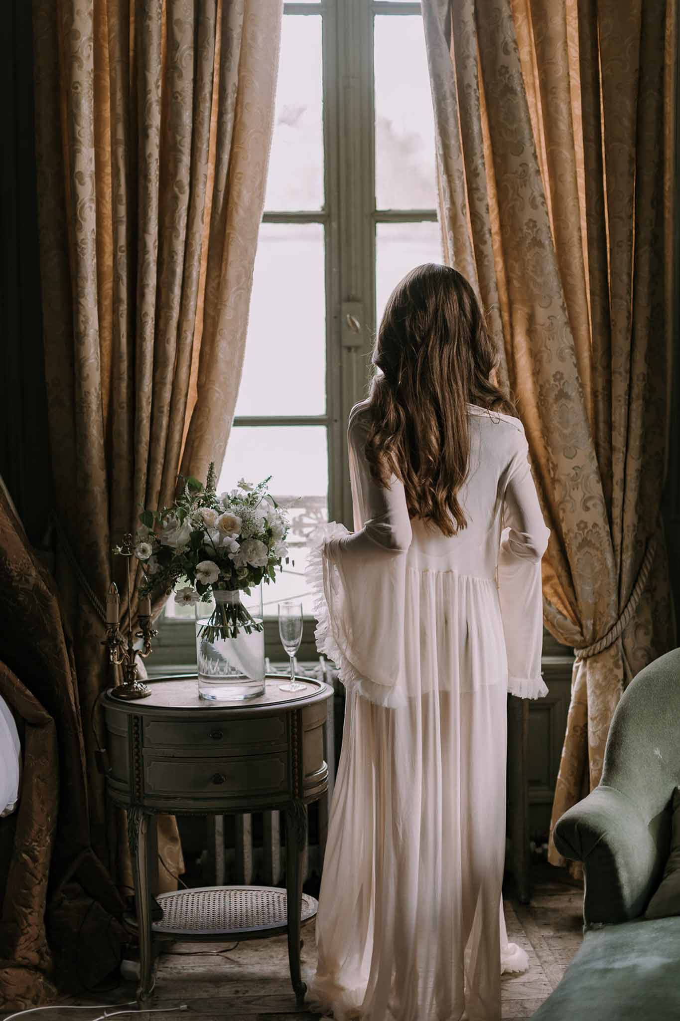 Bride from behind at window in ivory robe beside white and blush bouquet in chateau room with gold curtains