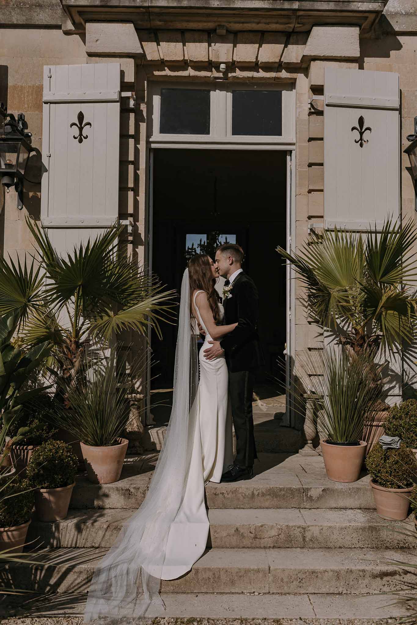 Couple nearly kissing at chateau doorway with fleur-de-lis shutters, cathedral veil trailing down stone steps