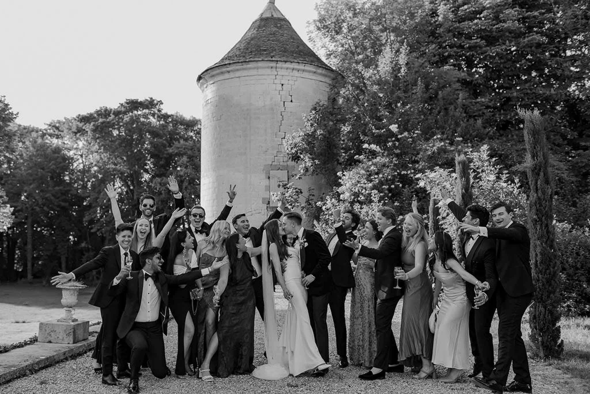 Black and white group of 16 guests cheering around kissing couple before round chateau stone tower