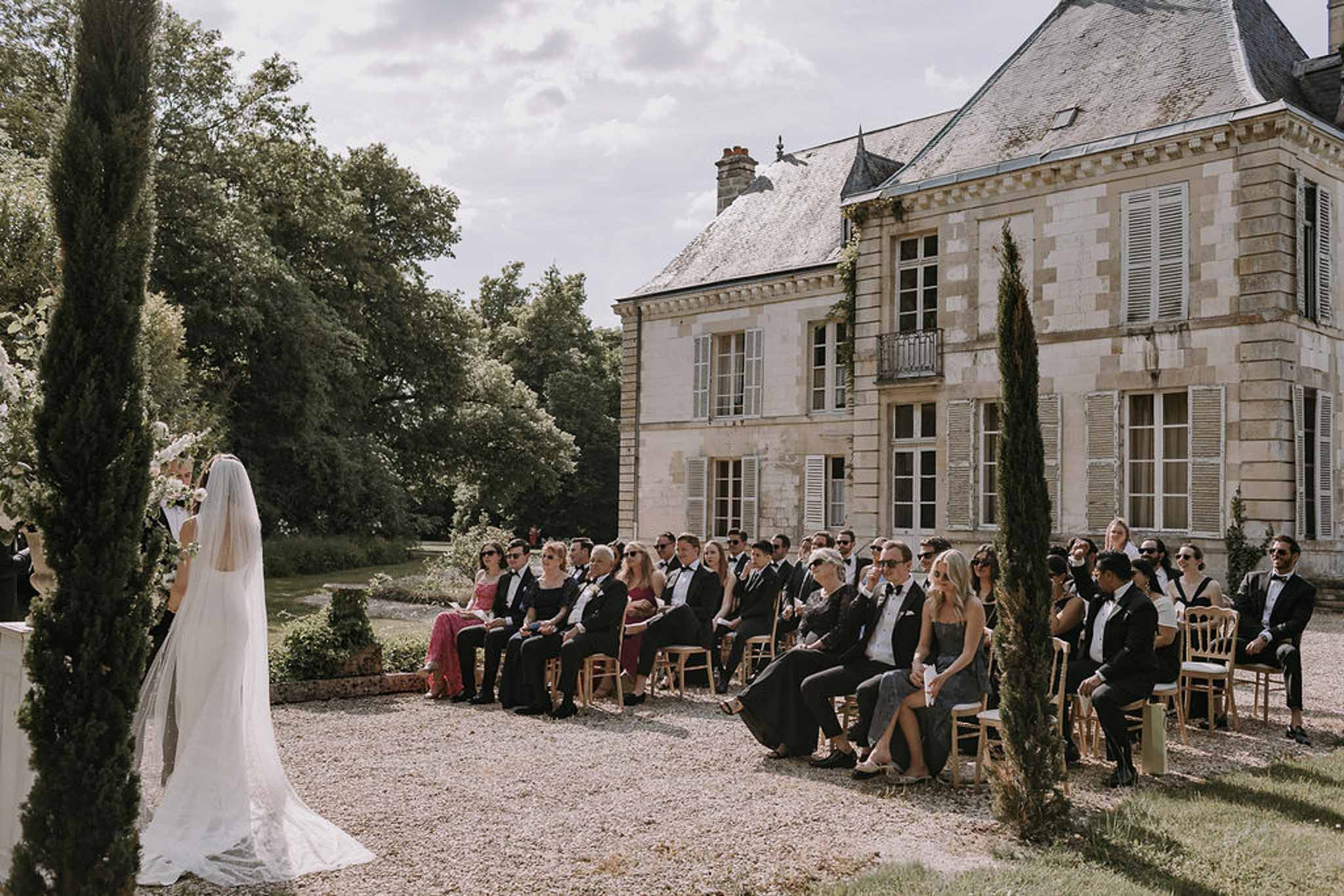 Bride at altar with cathedral veil as 45 guests sit in gold chiavari chairs before stone chateau