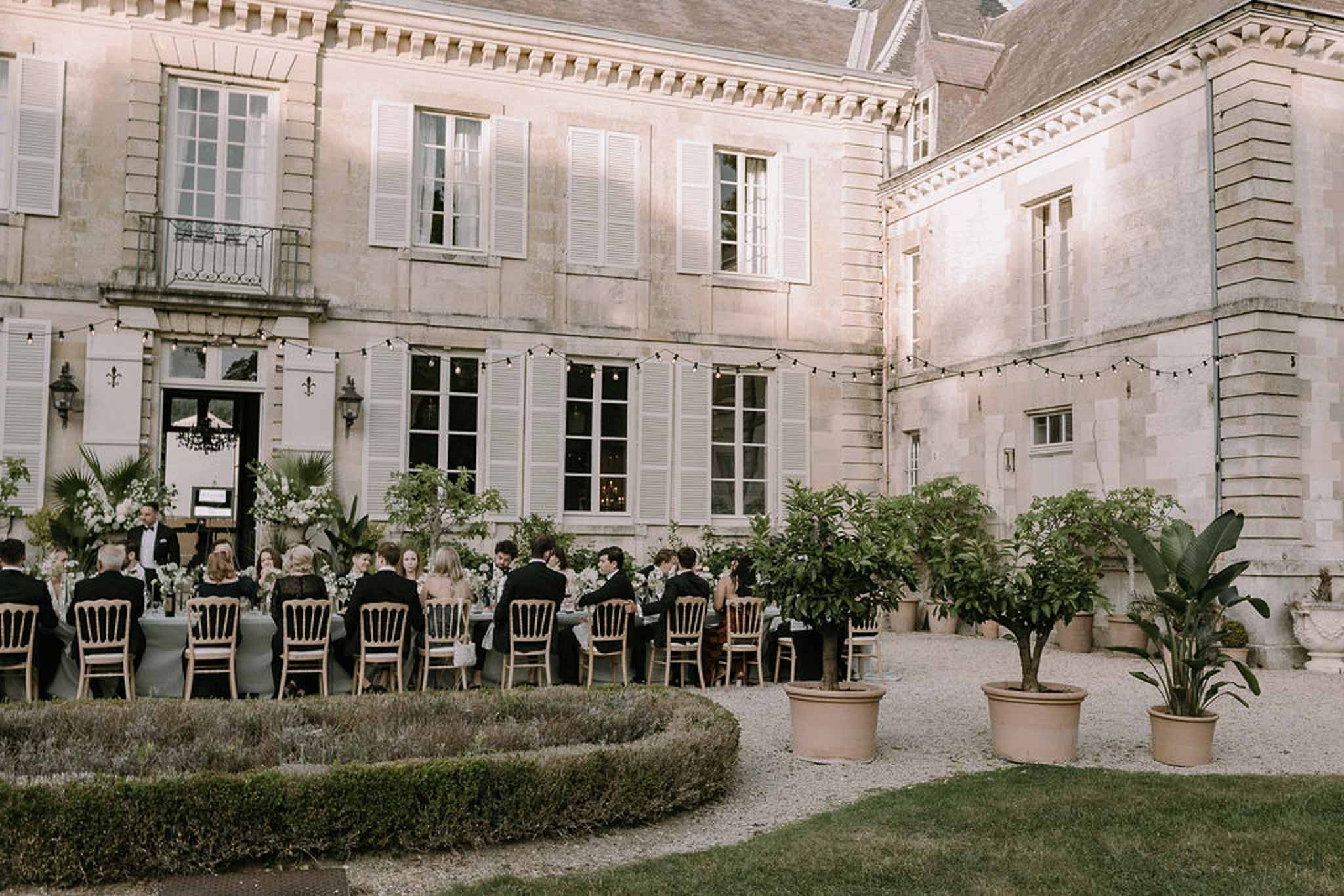 Twenty-five guests at slate-linened table with Napoleon chairs string lights and citrus trees before chateau at dusk