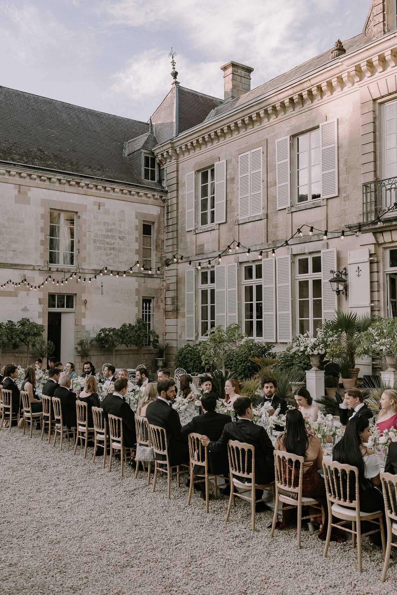 Forty guests at long candlelit table with white roses under string lights in chateau courtyard at dusk