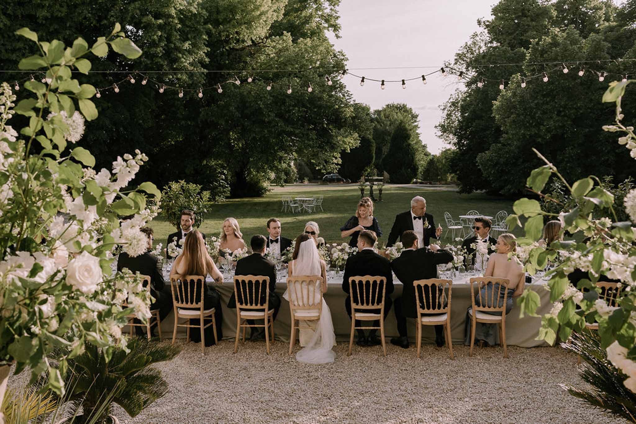 Long table with white rose runner and Edison lights as guest toasts bride and groom on terrace