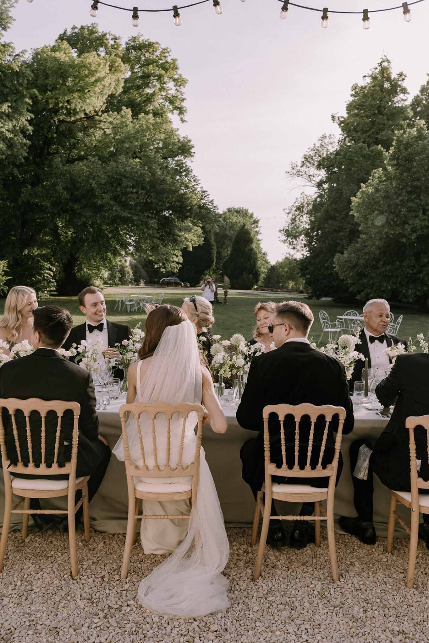 Intimate garden dinner with couple and eight guests at table with white allium bud vases under Edison lights