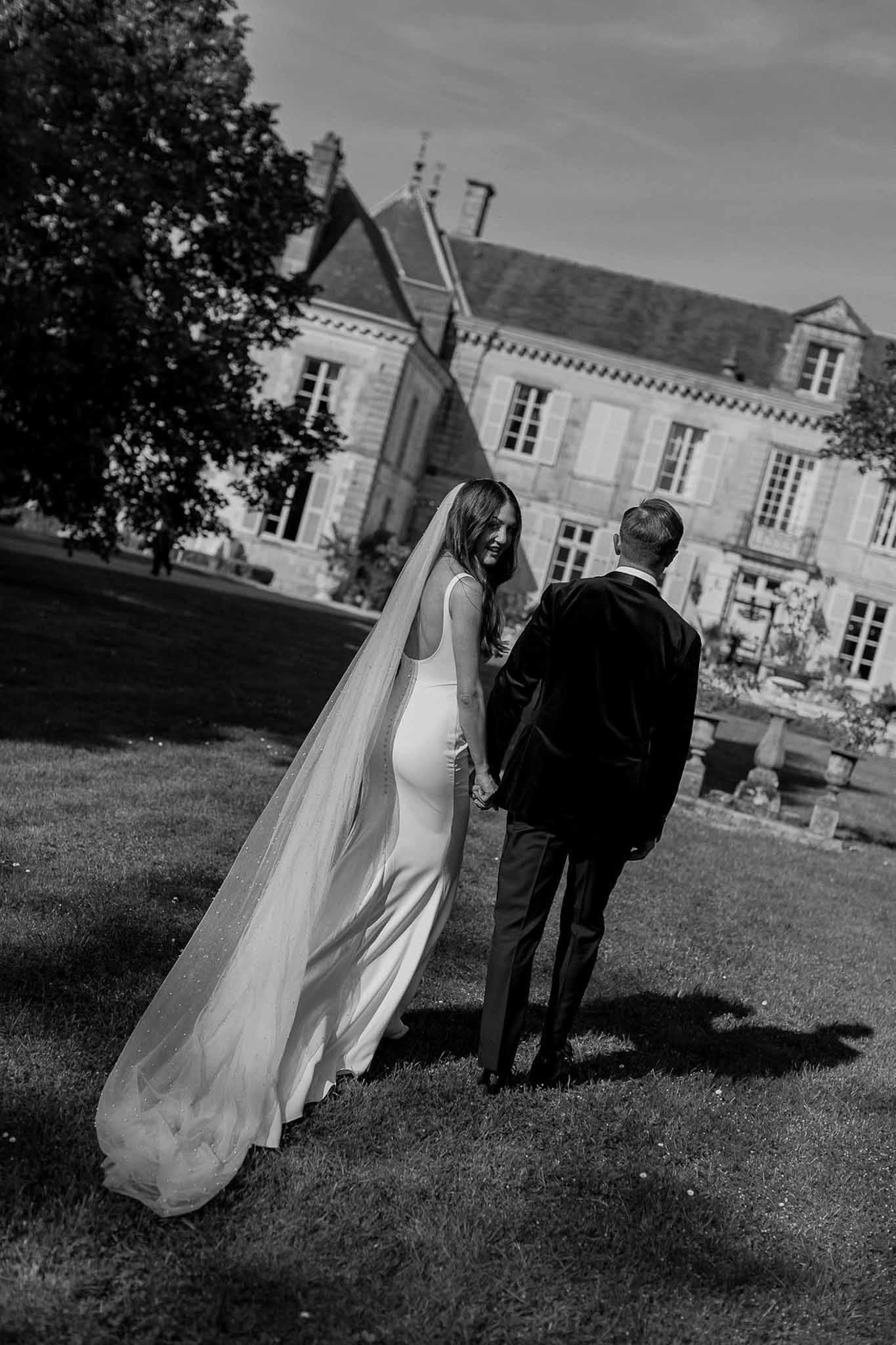Black and white rear view of couple walking across lawn with dotted cathedral veil trailing on grass