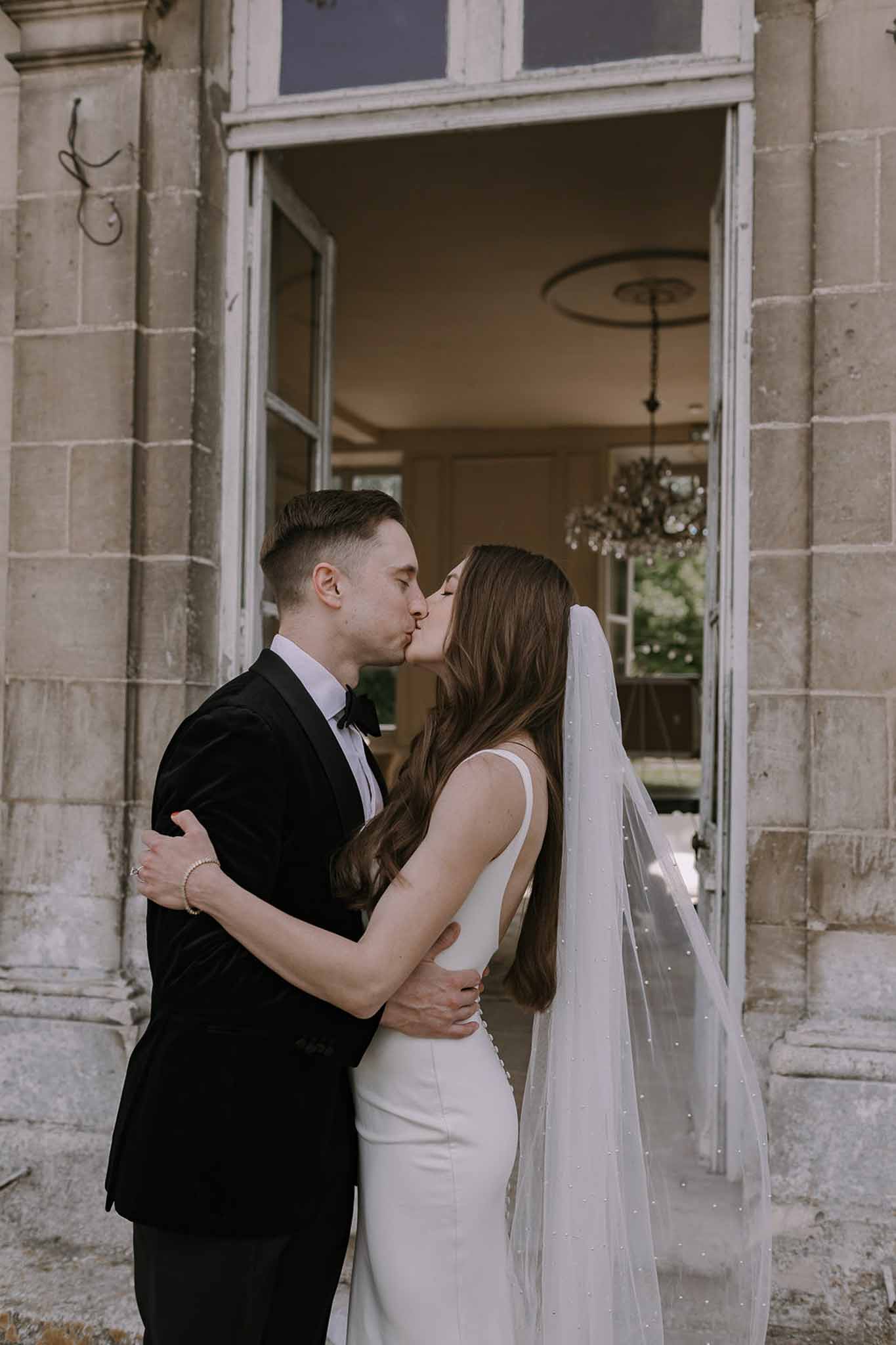 Couple kissing in chateau doorway with crystal chandelier, bride wearing pearl-embellished veil
