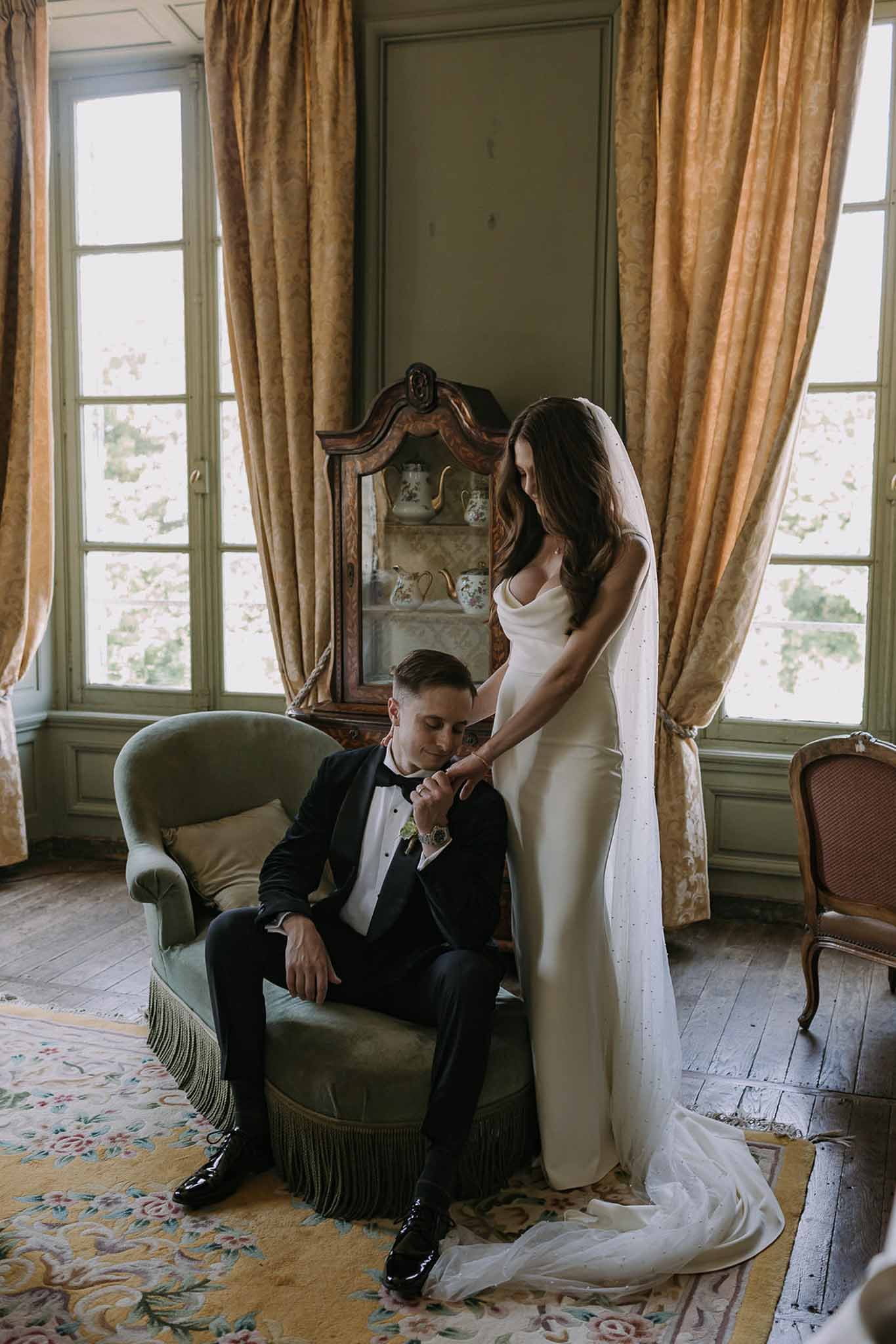 Groom in tuxedo kissing bride's hand as she stands in cathedral veil in sage-panelled chateau salon